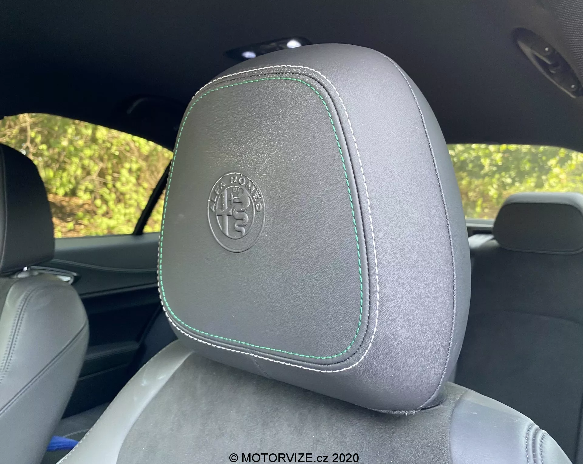 Gros plan sur un appui-tête de voiture dans l'Alfa Romeo Giulia 2016, avec une sellerie en cuir gris clair et des coutures contrastantes vert clair, vu depuis le siège arrière. L'appui-tête comporte un logo circulaire en relief suggérant une finition premium ou personnalisée. L'arrière-plan montre un feuillage flou à travers les vitres de la voiture, suggérant un cadre extérieur en plein jour.