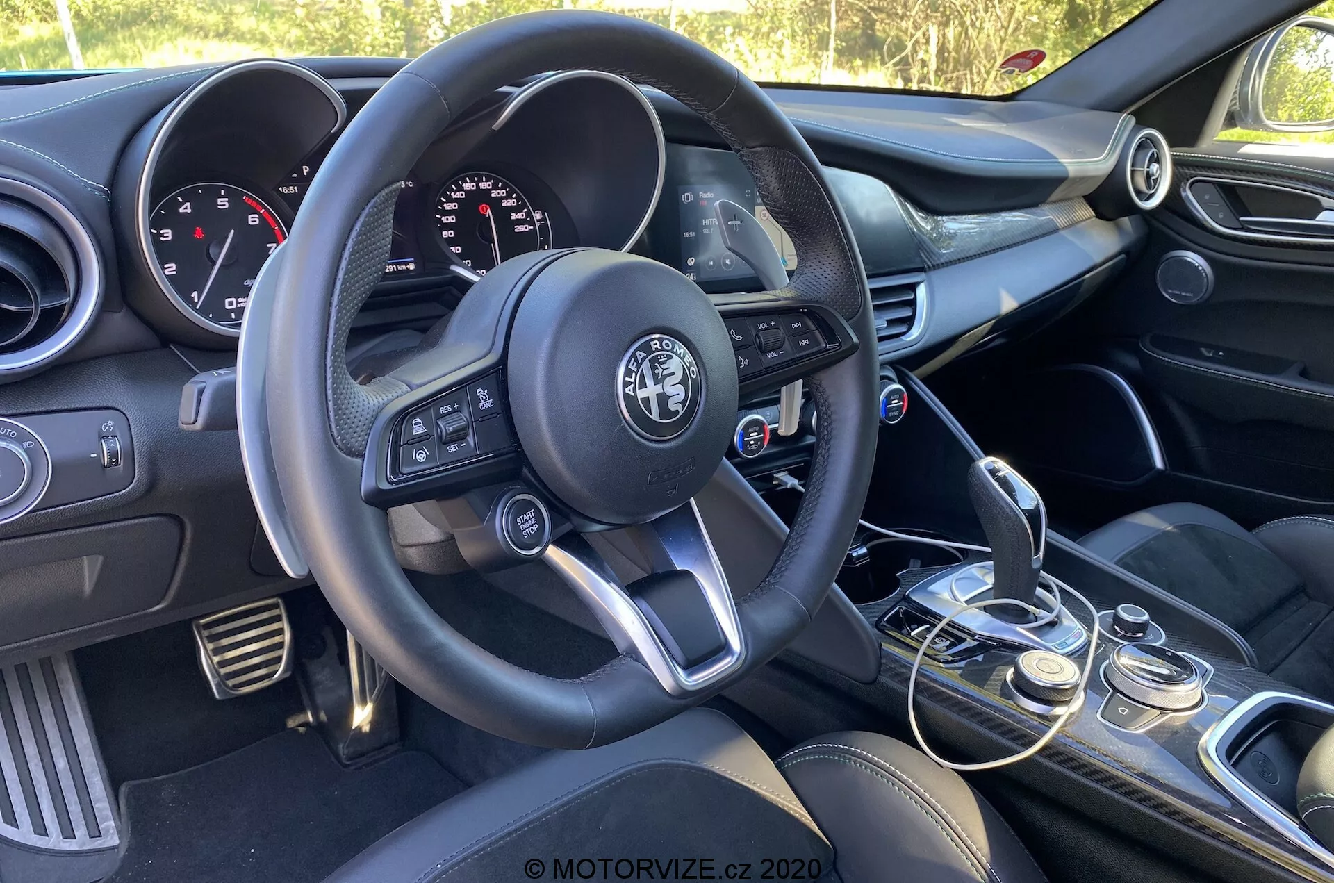 Vue intérieure de l'Alfa Romeo Giulia 2016 se concentrant sur le côté conducteur depuis la perspective du passager, montrant le volant à trois branches avec boutons multifonctions, le logo du constructeur bien en évidence, le tableau de bord avec cadrans analogiques et écran numérique, la console centrale avec commandes audio et climatisation, le levier de vitesse manuel avec souflet en cuir, le sélecteur de mode de conduite, les sièges avant avec rembourrage foncé, et le panneau de porte du conducteur partiellement visible avec commandes pour fenêtres et rétroviseurs.