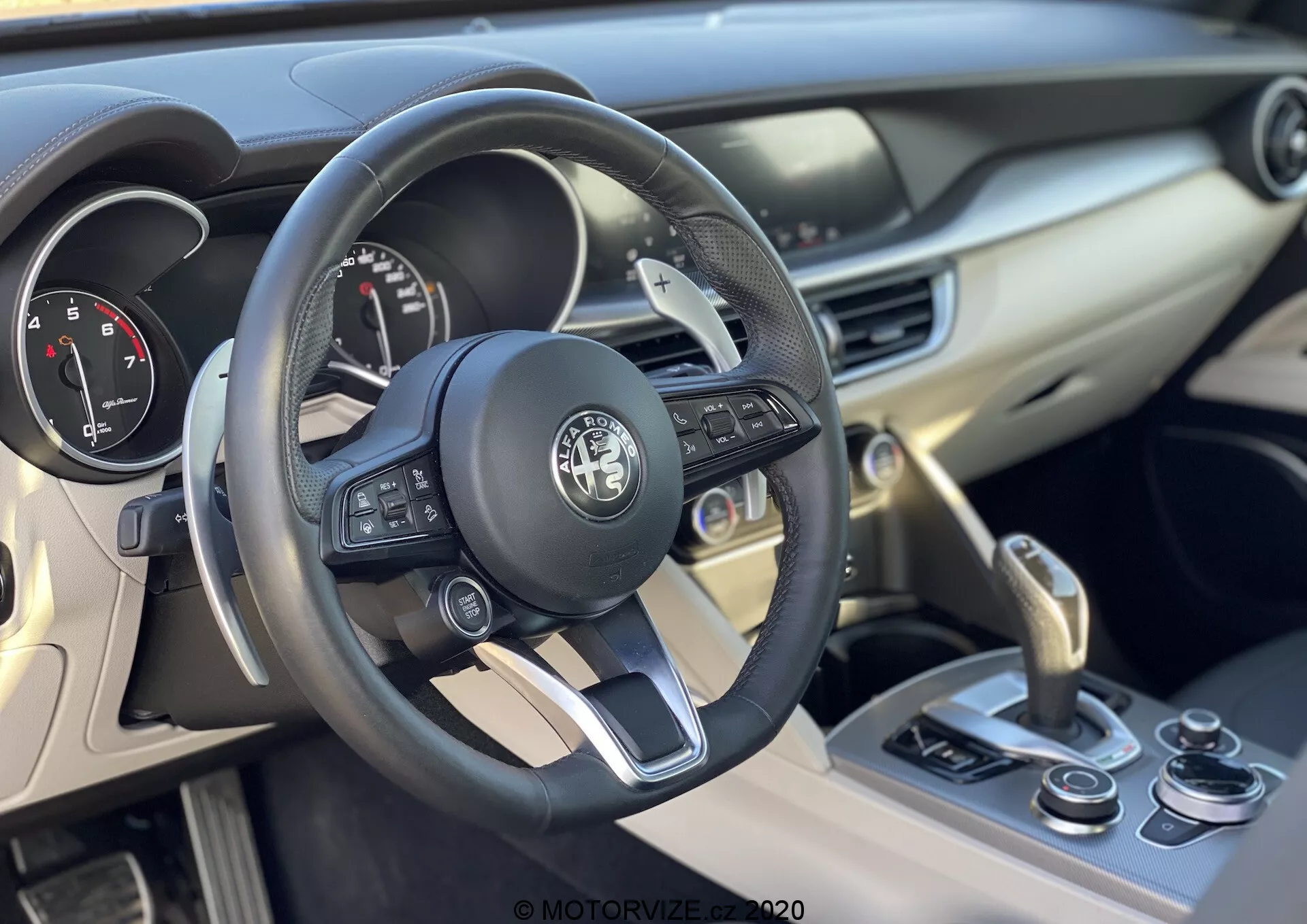 Vue intérieure d'une Alfa Romeo Stelvio 2017, axée sur le volant et le tableau de bord. Le volant en cuir avec des boutons multifonctions pour multimédia, appels et régulateur de vitesse présente l'emblème du fabricant au centre. Le tableau de bord derrière le volant comprend des cadrans analogiques pour le compteur de vitesse et le compte-tours, probablement avec un petit écran numérique pour des informations supplémentaires. La console centrale à droite comprend des bouches d'aération, un bouton de démarrage-arrêt du moteur et un levier de vitesses indiquant une transmission automatique, avec des commandes supplémentaires comme un sélecteur de modes de conduite. L'intérieur, avec des matériaux tels que des plastiques doux au toucher et du cuir avec des accents métalliques, suggère une attention particulière à l'ergonomie et au confort du conducteur.