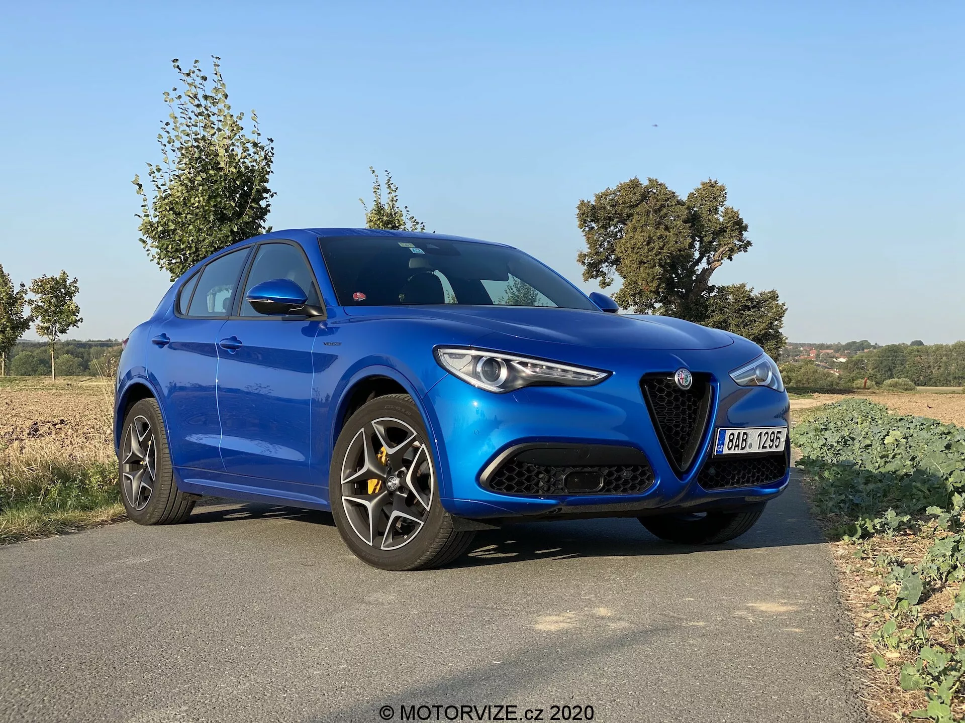 L'image montre une berline sportive Alfa Romeo Stelvio bleue de 2017 garée sur le bord de la route dans un environnement rural ou semi-rural. La voiture est inclinée, offrant une vue aux trois quarts de son avant et de son côté gauche, mettant en valeur sa calandre distinctive et ses phares. L'éclairage moderne et élégant, le design du capot profilé, les jantes en alliage noires à rayons multiples et les étriers de frein orientés performance sont tous visibles. L'éclairage naturel indique que la photo a été prise en journée, probablement le matin ou en fin d'après-midi, avec des arbres et des champs ouverts en arrière-plan offrant une atmosphère sereine.
