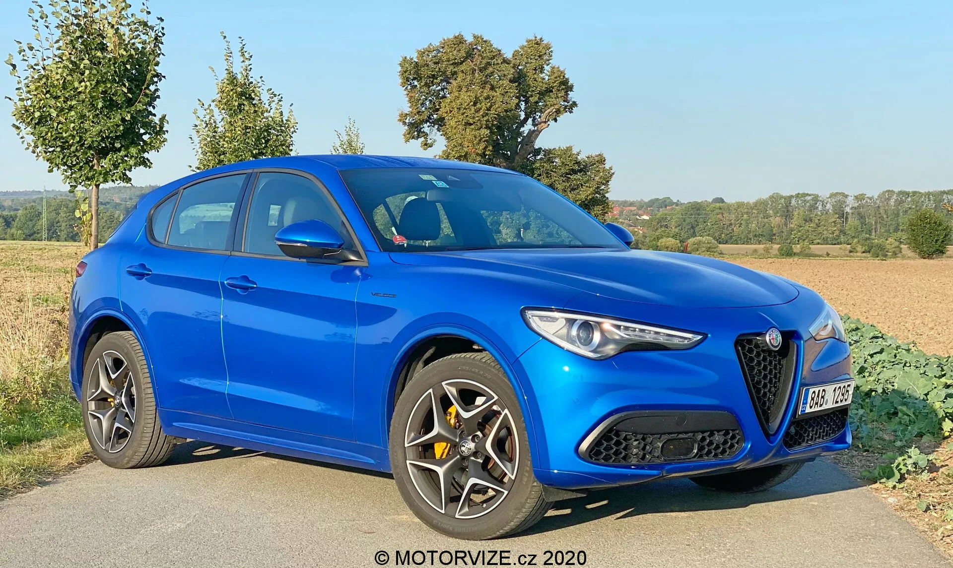 Un SUV bleu, photographié sous un angle trois-quarts avant, mettant en avant les profils avant et latéral. Capturée en pleine journée sous un ciel dégagé, cette image met en valeur les caractéristiques de design du véhicule, dont une calandre avant distinctive avec un emblème, des phares LED énergiques, et de larges prises d'air dans le pare-chocs avant. Les lignes sculptées du capot ajoutent une esthétique sportive. Le profil latéral dévoile de grandes roues en alliage à plusieurs branches, une ligne de toit inclinée, des portes latérales sculptées, des rétroviseurs latéraux avec clignotants intégrés et des garnitures de fenêtre. Les caractéristiques technologiques visibles incluent les phares LED, les feux de jour, les poignées de portes couleur carrosserie avec entrée sans clé et les rétroviseurs latéraux réglables électriquement. Le véhicule est stationnaire sur le côté droit d'une route ou d'un sentier avec de la végétation environnante, suggérant un cadre rural. Avec un filigrane indiquant l'année 2020, l'image est claire et bien focalisée.