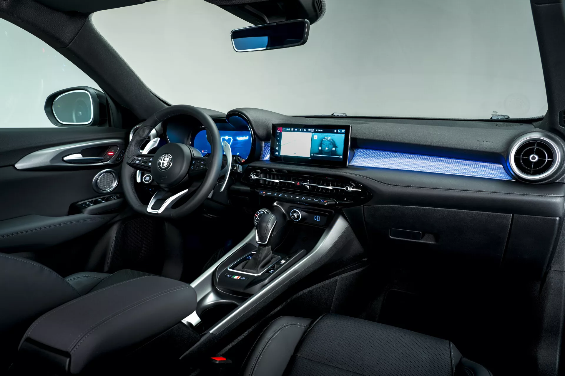 Vue intérieure du côté passager de l'Alfa Romeo Tonale 2022, mettant en évidence le volant multifonction, le tableau de bord numérique, le levier de vitesses automatique avec soufflet en cuir, le sélecteur de mode de conduite, le système d'infodivertissement avec écran, l'éclairage ambiant et les sièges en cuir premium avec coutures contrastées.