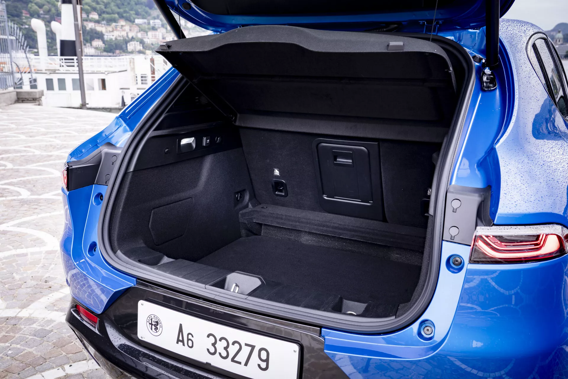 Vue angulaire rapprochée de l'arrière de l'Alfa Romeo Tonale 2022 bleu présentant l'espace du coffre ouvert avec un intérieur moquetté, des compartiments intégrés et une prise de puissance, à côté du pare-chocs arrière visible avec des capteurs de stationnement et des feux arrière LED modernes.