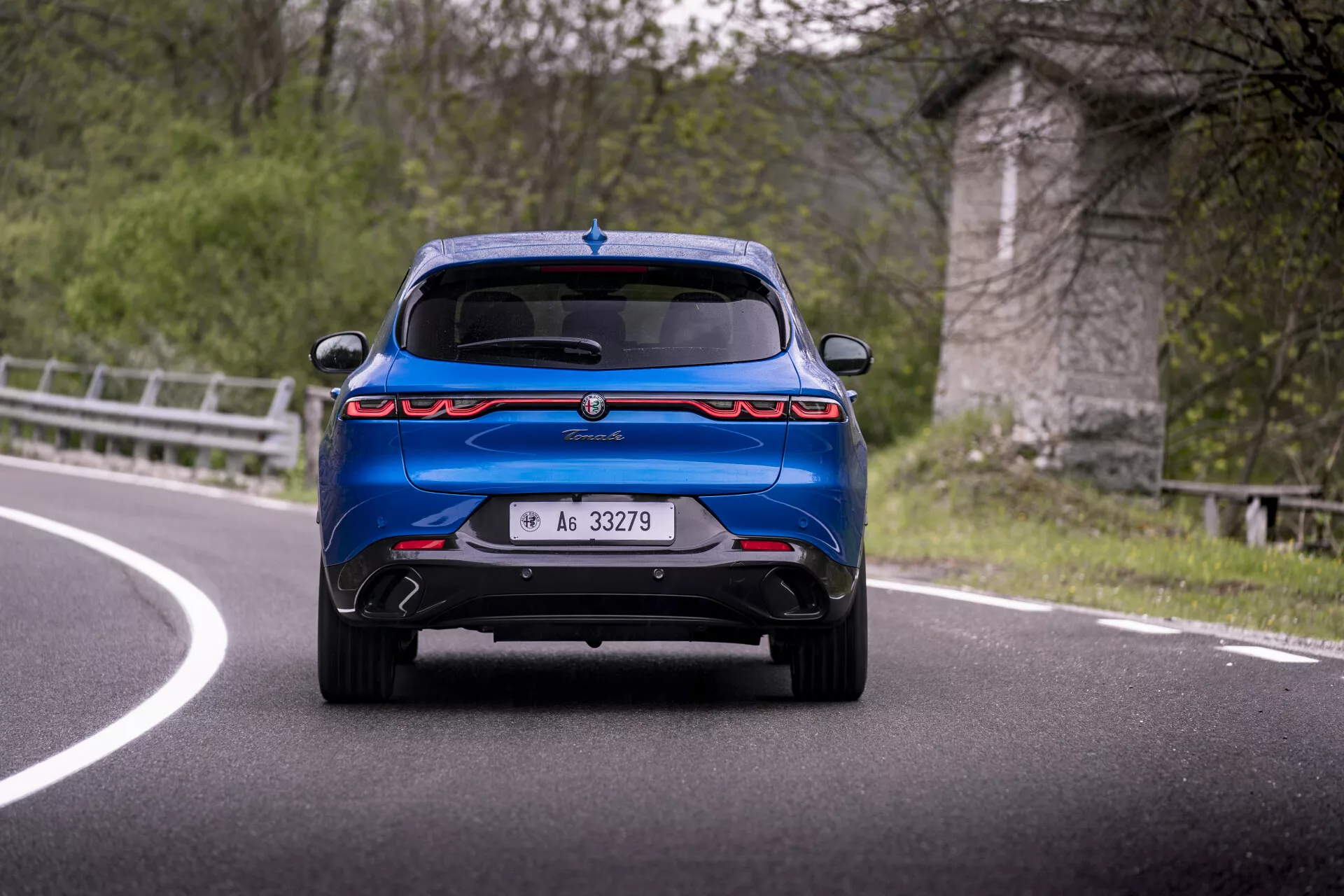 Alfa Romeo Tonale 2022 capturé en mouvement sur une courbe, montrant sa vue arrière avec des feux arrière horizontaux distinctifs, un pare-chocs sportif avec double échappement, spoiler et capteurs de stationnement contre un arrière-plan pittoresque