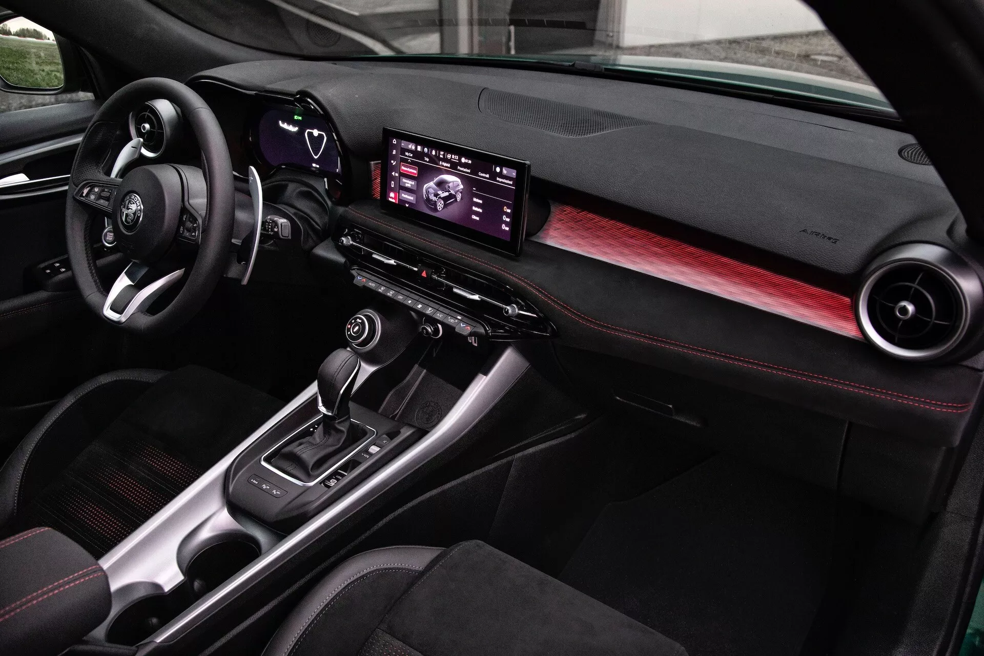 Intérieur de l'Alfa Romeo Tonale 2022 montrant un tableau de bord moderne, un centre d'infodivertissement numérique, des accents en fibre de carbone et des sièges cousus en rouge, vu du côté passager avant