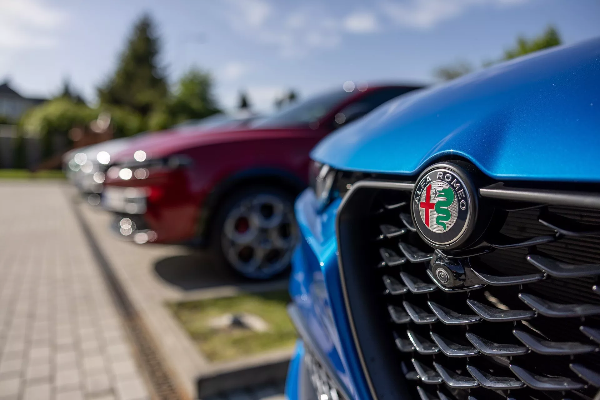 Vue rapprochée de la calandre avant de l'Alfa Romeo Tonale 2022 bleue, centrée sur l'emblème, avec des lamelles verticales et un arrière-plan flou d'autres véhicules