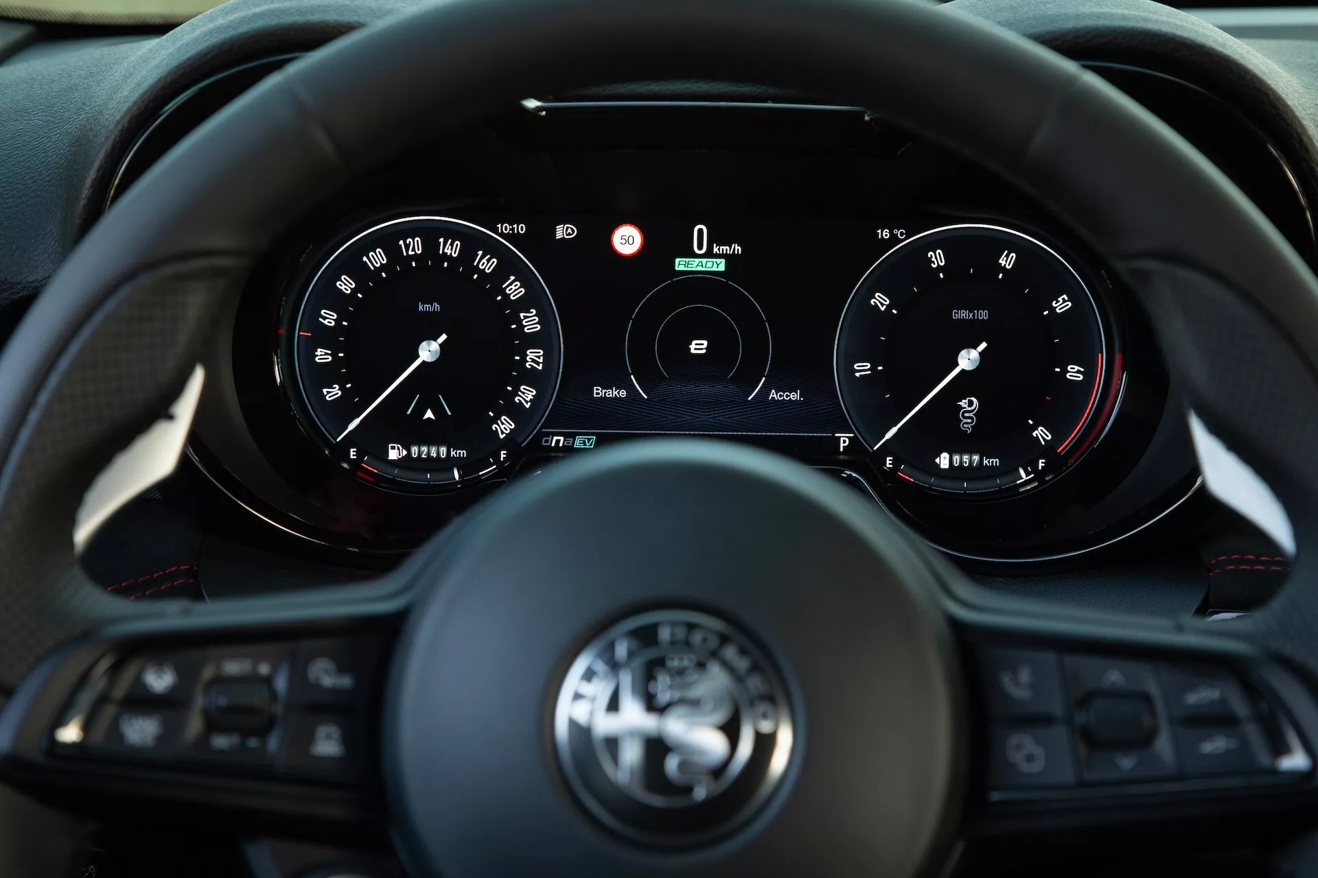 Intérieur de l'Alfa Romeo Tonale 2022 montrant la vue du conducteur sur un volant multifonction à trois branches et un cluster d'instruments avec des jauges analogiques et un affichage numérique central indiquant une vitesse et un RPM de zéro, caractérisant les technologies de connectivité et d'assistance au conducteur