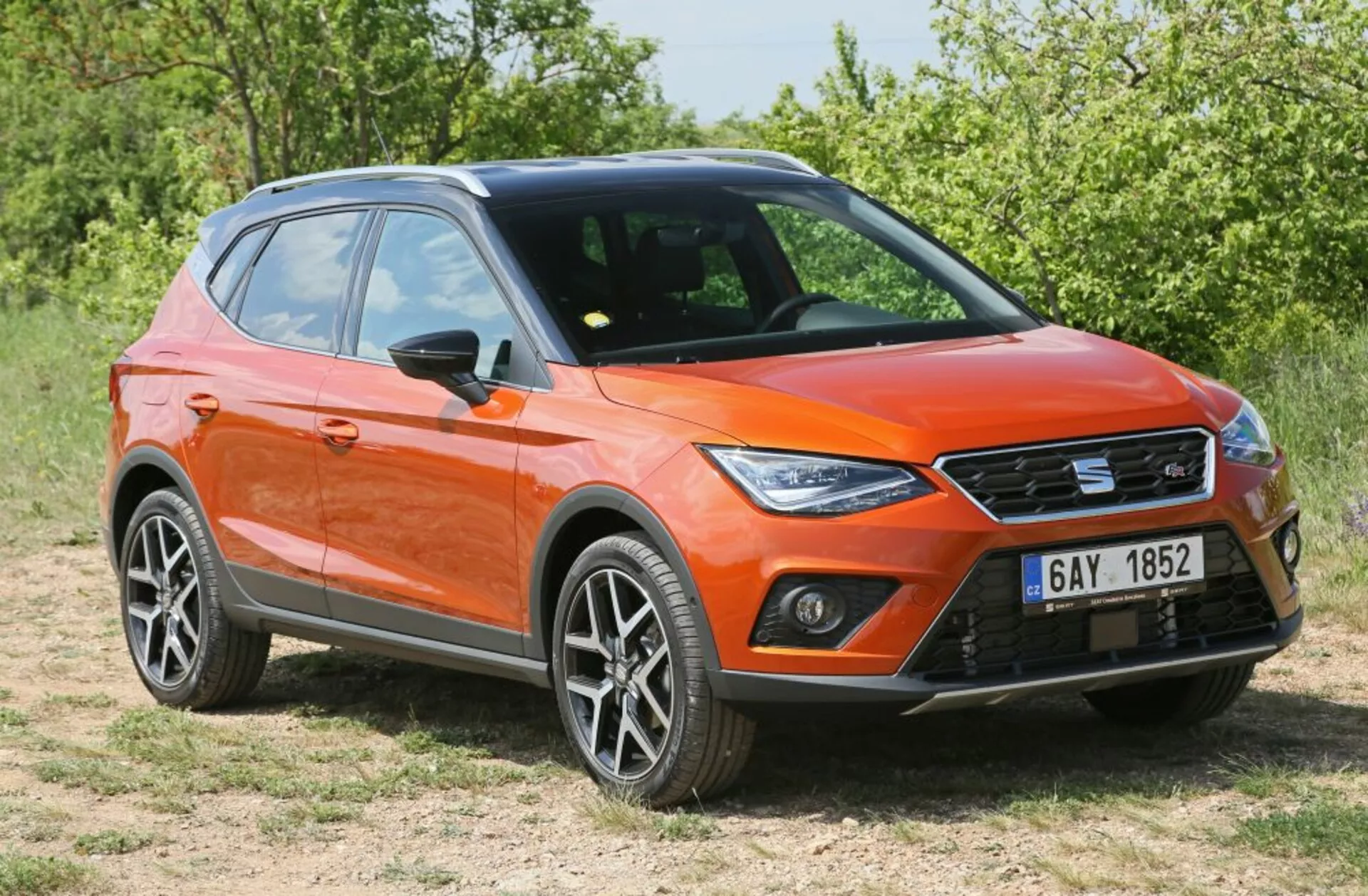 Crossover/SUV orange garé sur l'herbe dans une vue frontale trois quarts affichant des feux de jour LED, un design moderne et affûté avec des roues en alliage et des barres de toit sous la lumière du soleil