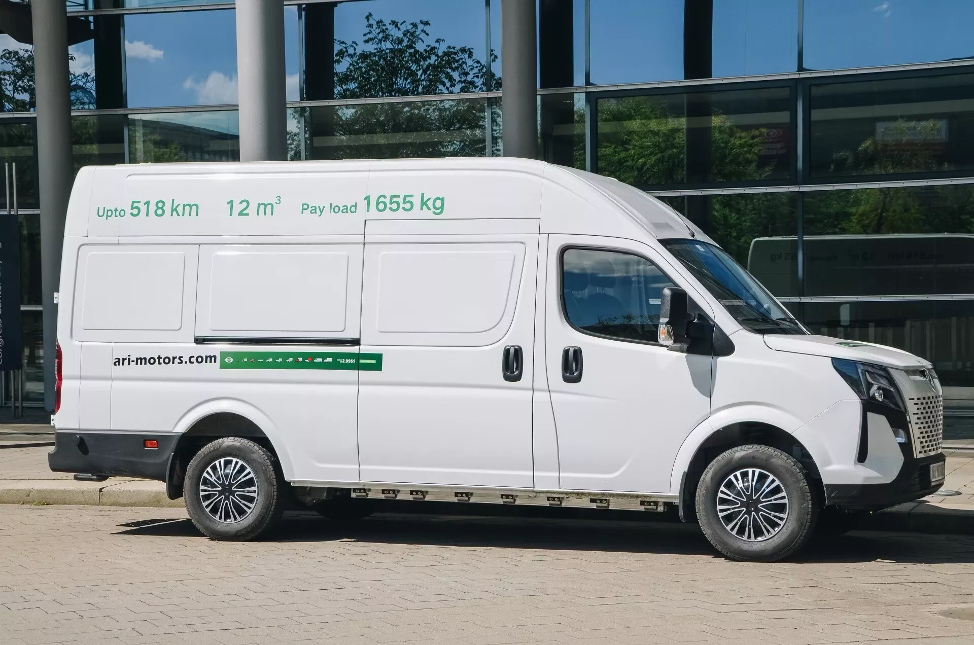Un fourgon commercial blanc, l'ari-motors 1710 2024 (1ère génération), est vu de profil latéral dans une photographie professionnelle et détaillée. L'image capture tout le côté passager, révélant une porte coulissante et deux fenêtres sur la moitié supérieure du véhicule. Le texte sur le côté du fourgon indique les caractéristiques clés : 'Jusqu'à 518 km' d'autonomie, 'Charge utile 1655 kg' et '12 m³' de volume de chargement. Cela suggère que le fourgon peut être électrique ou utiliser un carburant alternatif. Positionné contre un bâtiment aux murs de verre, la photo est prise à la lumière du jour avec des ombres sur la droite, soulignant le design moderne du fourgon et son usage commercial.