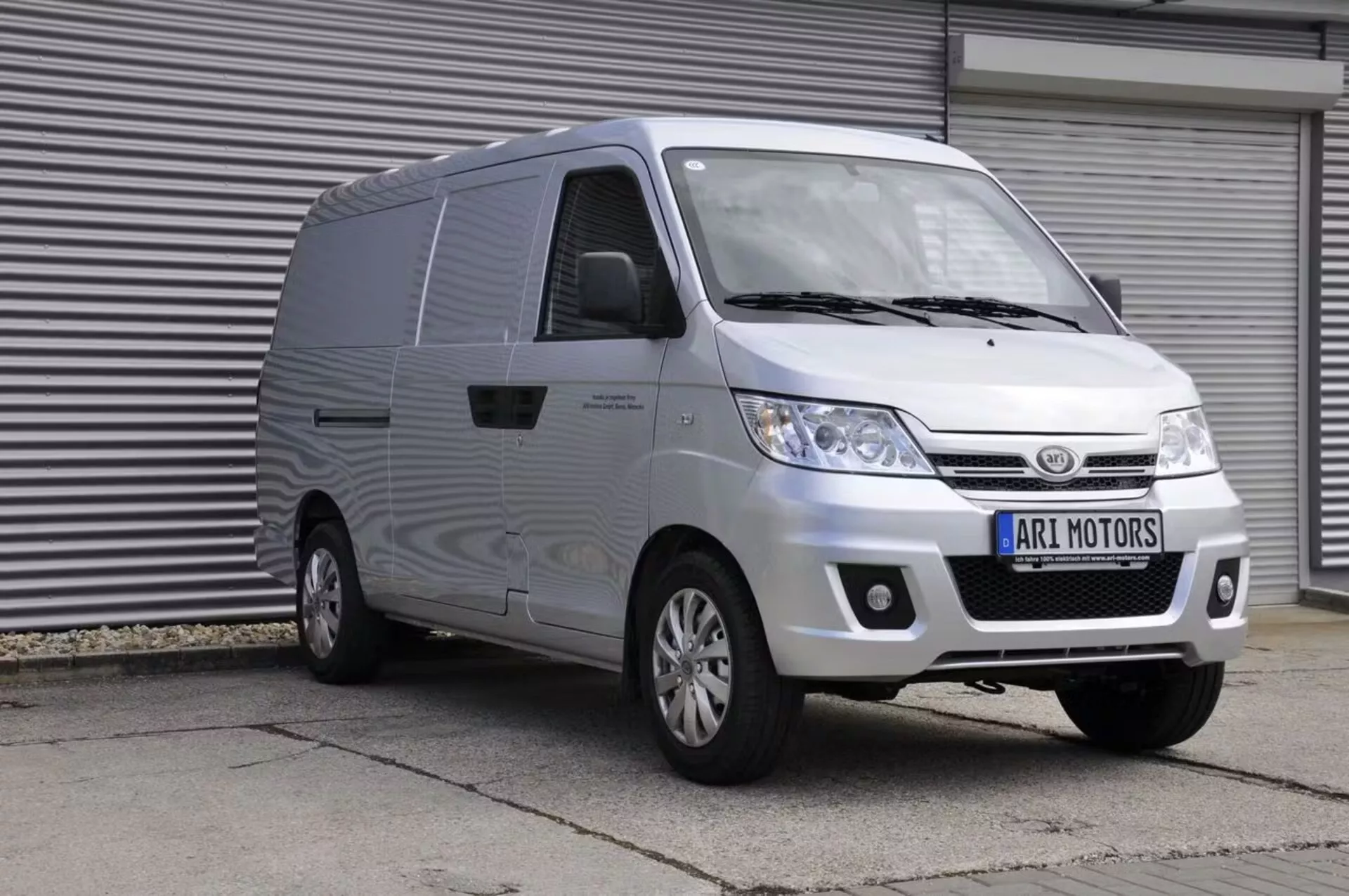 Image d'une fourgonnette blanche ARI Motors 901 2024 vue de trois quarts de face. La fourgonnette présente une calandre proéminente avec des lamelles horizontales, de grands phares et un pare-chocs avant avec d'éventuels feux de brouillard. Sur le côté, une porte coulissante du côté passager facilite l'accès à la zone de chargement. La fourgonnette dispose de rétroviseurs latéraux de taille adéquate et de jantes à rayons multiples. Photographiée d'une légère élévation, l'image offre une vue claire du capot, du pare-brise et du toit, et indique qu'elle est stationnée dans un environnement industriel avec des volets roulants en arrière-plan. Le texte 'ARI MOTORS' apparaît sur la plaque d'immatriculation.