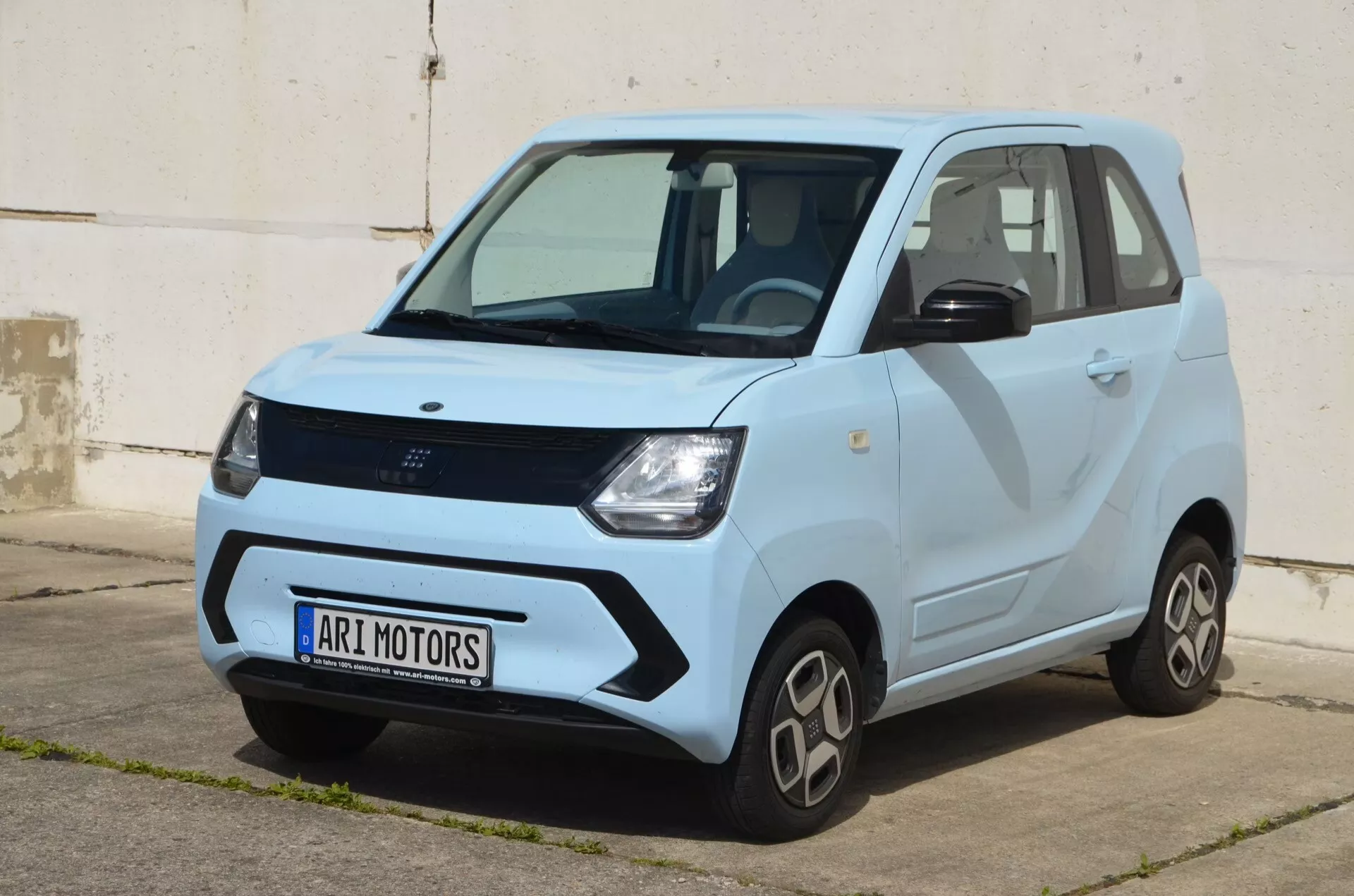 Vue extérieure de trois quarts avant de la Ari-Motors Soleno 2024, mettant en valeur son design électrique compact. L'image met en avant la zone de la calandre avant, les phares, la roue avant et le profil du conducteur à la lumière naturelle, révélant les lignes de carrosserie et un schéma de couleurs contrastantes avec un ton plus sombre sur la moitié inférieure. Les éléments de design pratiques tels que les passages de roue évasés, les roues de taille modeste et les poignées de porte distinctes sont visibles. Un pli proéminent s'étend de l'arc de roue avant à l'arrière, ajoutant du caractère. La petite taille indique une adaptation aux environnements urbains.