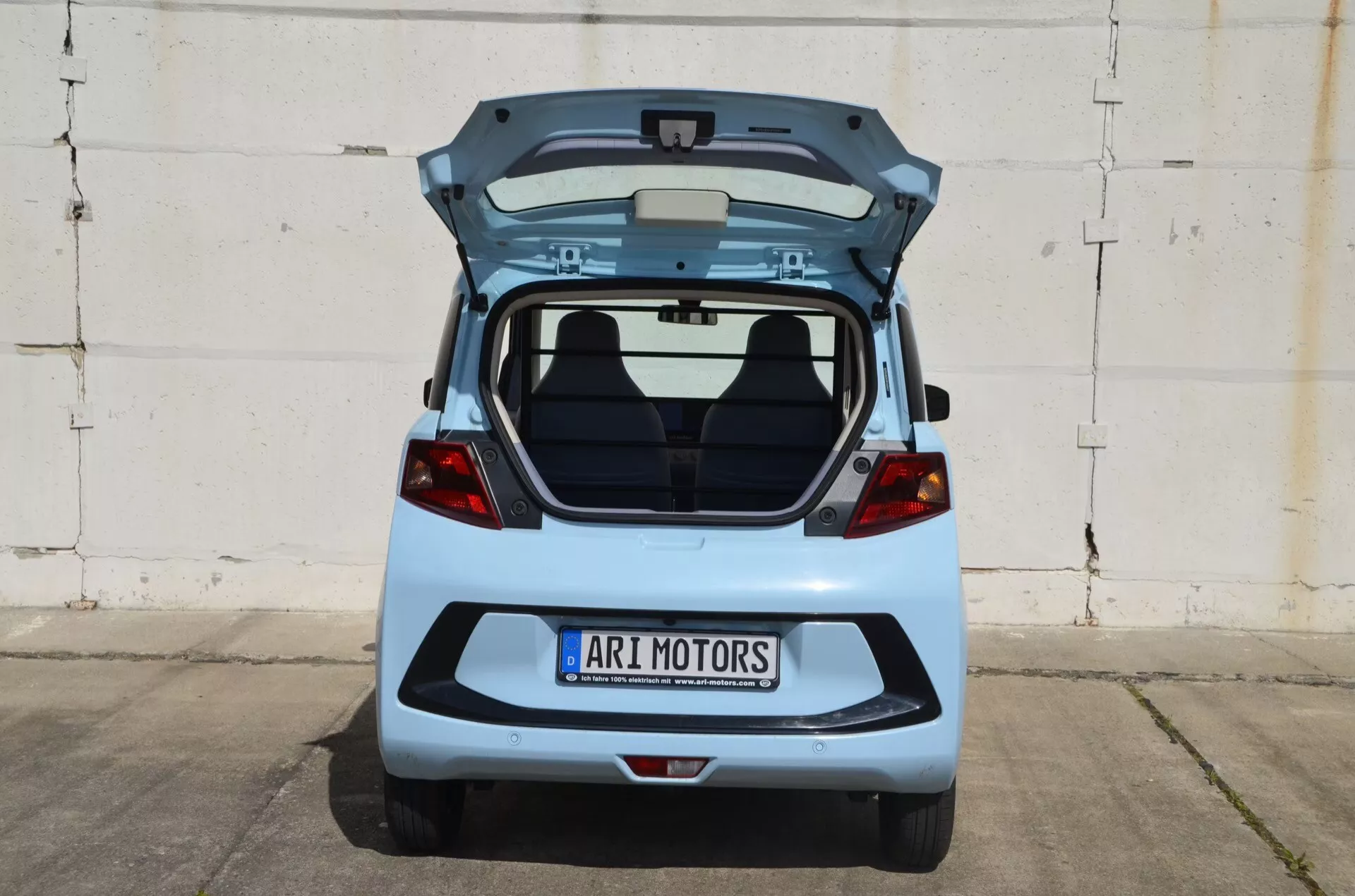 Vue arrière de l'ARI Motors Soleno 2024 (1ère génération) avec coffre ouvert, montrant l'espace de chargement, les sièges arrière et l'intérieur du couvercle. Plaque d'immatriculation centrale avec badge 'ARI MOTORS' au-dessus. Peinture argentée/bleu claire et brillante reflétant l'environnement, prise de vue symétrique mettant en valeur le design arrière sans fonctionnalités technologiques visibles.