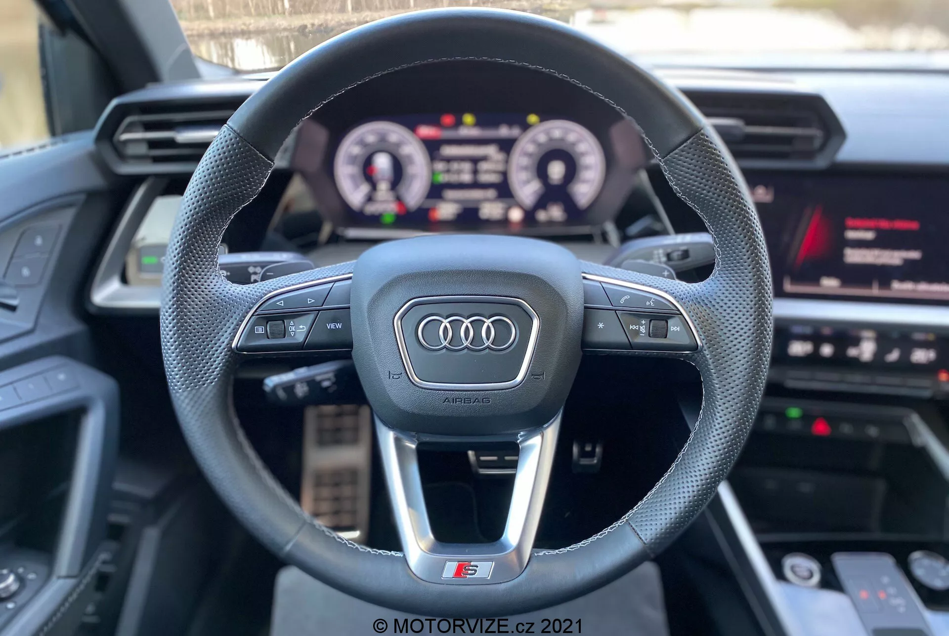 Vue à la première personne depuis le siège du conducteur d'une Audi A3 2020 (A3 Sportback) montrant le volant sportif à trois branches avec le logo Audi, des boutons de commande multifonctionnels, un tableau de bord numérique illuminé et une partie du système d'infodivertissement avec médias, navigation et réglages du véhicule. Les caractéristiques mettent en avant la technologie interactive et les composants de luxe avec un accent sur l'expérience de conduite.