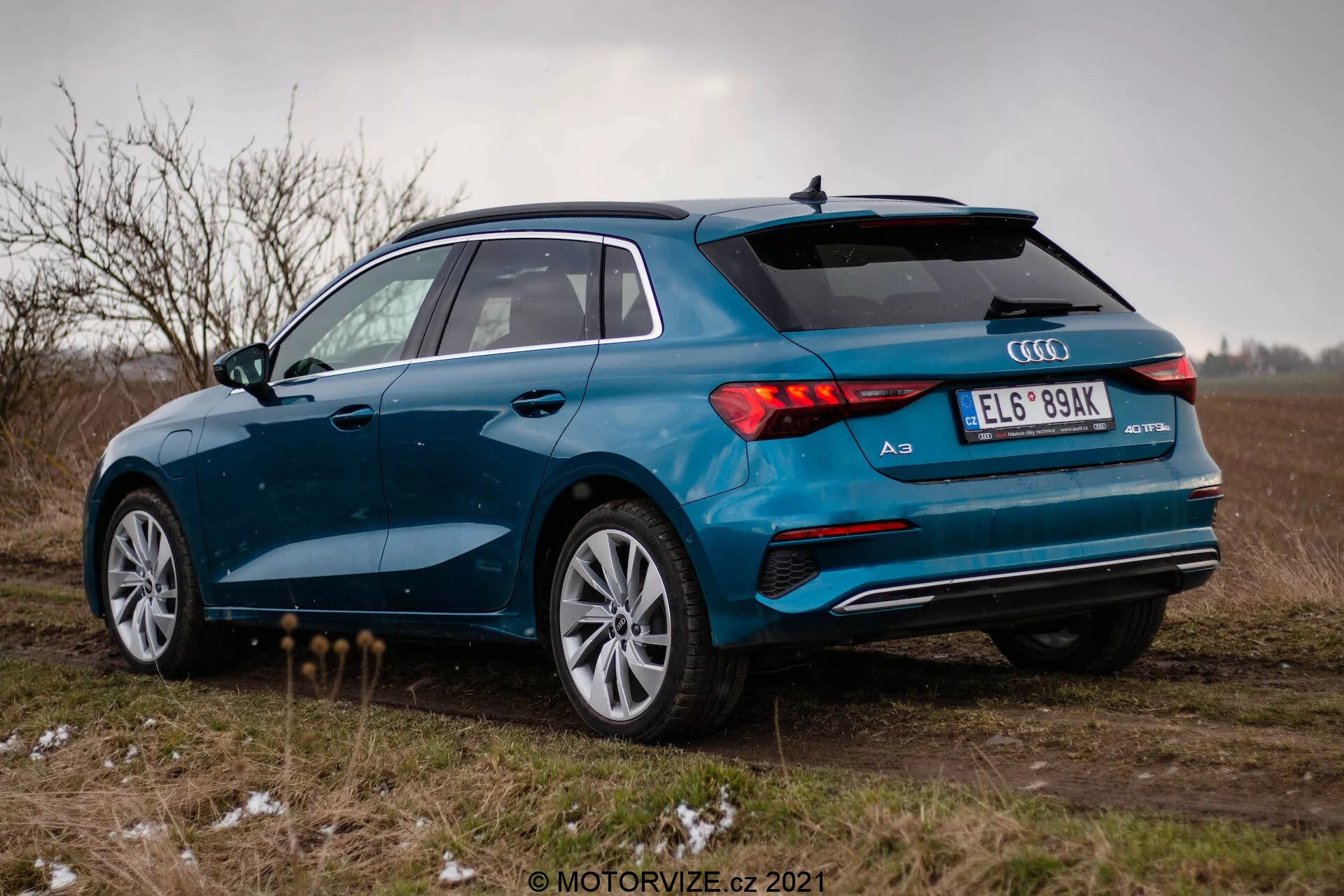 Une Audi A3 2020 (Sportback) bleue photographiée sous un angle trois-quarts arrière dans un champ ouvert sous un ciel nuageux. L'image met en évidence le côté droit de la voiture, les feux arrière à LED, le système d'échappement double, les jantes en alliage à plusieurs rayons et le spoiler arrière. Le véhicule est garé sur une surface non goudronnée avec des touffes d'herbe et une touche de neige ou de givre, suggérant des conditions météorologiques froides. L'ambiance globale est sombre avec un accent sur le design sportif et premium de l'Audi A3.