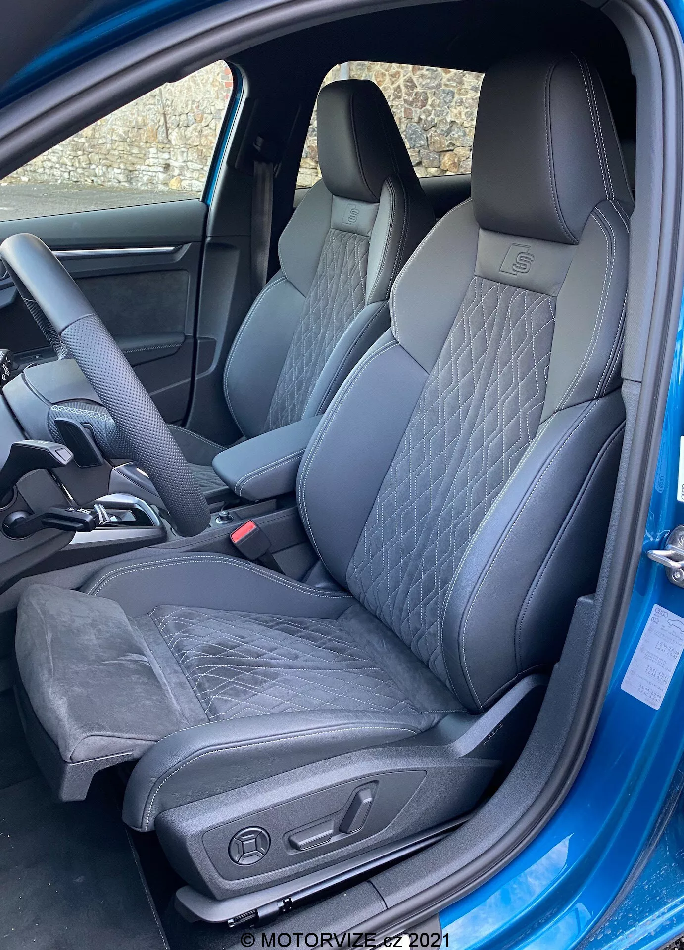 Vue extérieure à mi-angle de l'intérieur de l'Audi A3 Sportback 2020, focalisée sur le siège du conducteur. La porte est complètement ouverte, révélant un siège baquet sportif et moderne en cuir et possiblement Alcantara. Le siège présente des motifs de couture matelassée et des renforts latéraux prononcés pour le soutien latéral. Les commandes de réglage électrique du siège sont visibles, suggérant des ajustements de hauteur, d'inclinaison et de soutien lombaire. L'intérieur est monochromatique avec des tons sombres, contribuant à une sensation premium. Une partie du volant ergonomique avec un repose-pouce est également visible, indiquant un volant sportif.