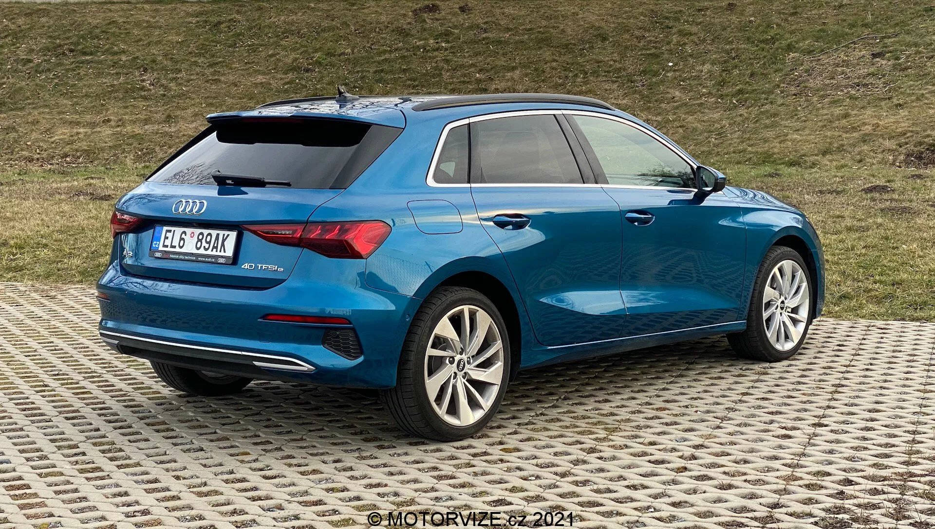 L'image montre une Audi A3 2020 Sportback bleue vue de trois quarts arrière, mettant en évidence le côté arrière droit. Les détails clés incluent des jantes en alliage multi-branches avec une finition argentée, des feux arrière LED partiellement visibles, une carrosserie aérodynamique avec une ligne d'épaule prononcée, des badges à l'arrière indiquant le modèle et peut-être la version, des vitres arrière teintées pour plus d'intimité, un petit spoiler de toit, des barres de toit pour la polyvalence et des doubles sorties d'échappement intégrées dans le pare-chocs arrière inférieur. La voiture est garée sur une surface à motifs avec un fond herbeux, suggérant un environnement pas trop urbain.