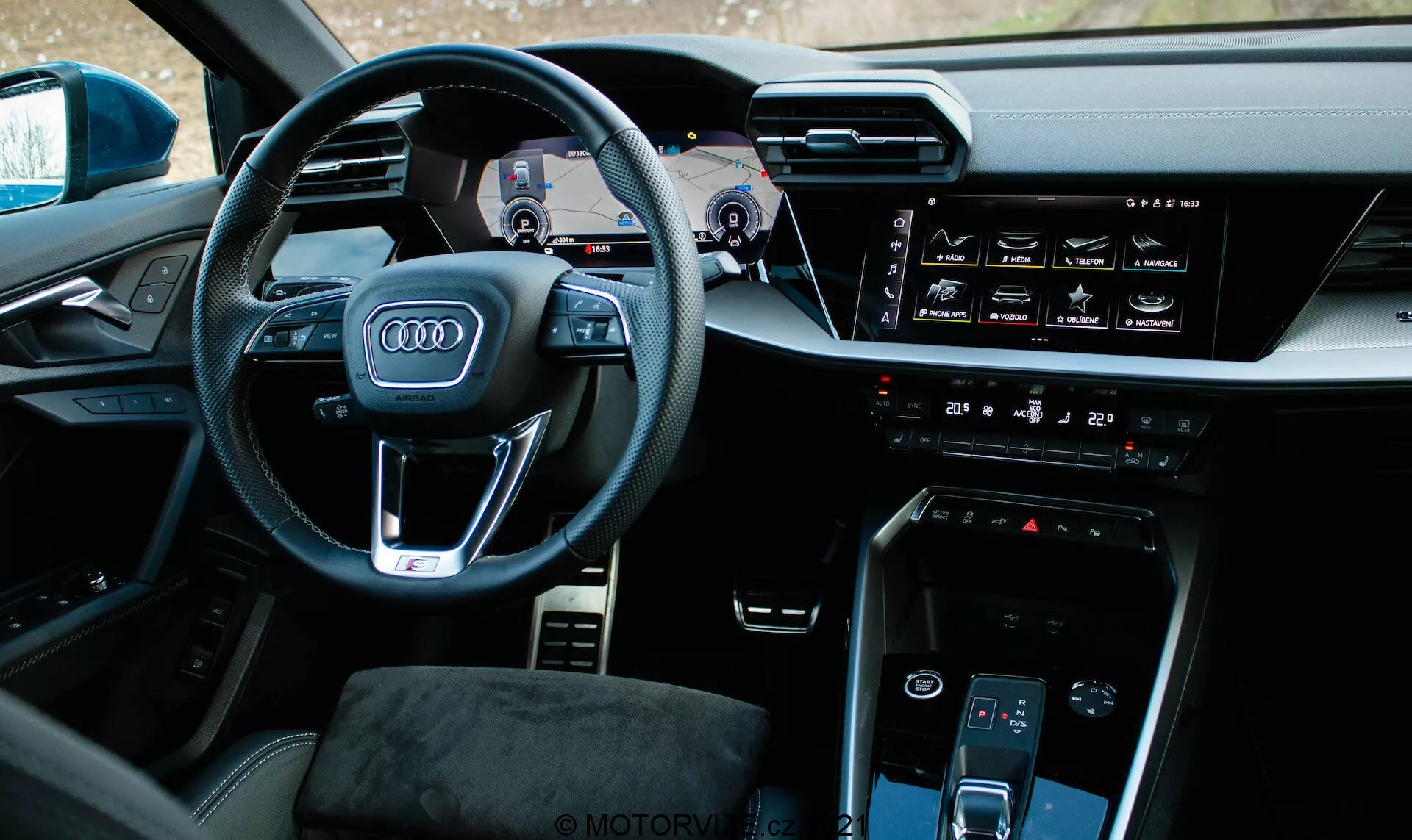 Vue intérieure d'une Audi A3 Sportback 2020 depuis un angle légèrement désaxé à gauche, centrée sur le côté conducteur. Le volant proéminent arbore le logo emblématique des quatre anneaux d'Audi, des boutons multifonctions et des palettes de changement de vitesse visibles. Derrière le volant, le tableau de bord numérique connu sous le nom d'Audi Virtual Cockpit affiche le compteur de vitesse, le compte-tours et la carte de navigation. La console centrale comprend un écran tactile horizontal pour l'infodivertissement, une interface de commande de climatisation, un bouton de démarrage-arrêt du moteur, un autre écran tactile pour la commande de climatisation et un sélecteur de vitesses minimaliste. L'intérieur présente des matériaux de haute qualité et un design technologiquement intégré avec une sensation premium.