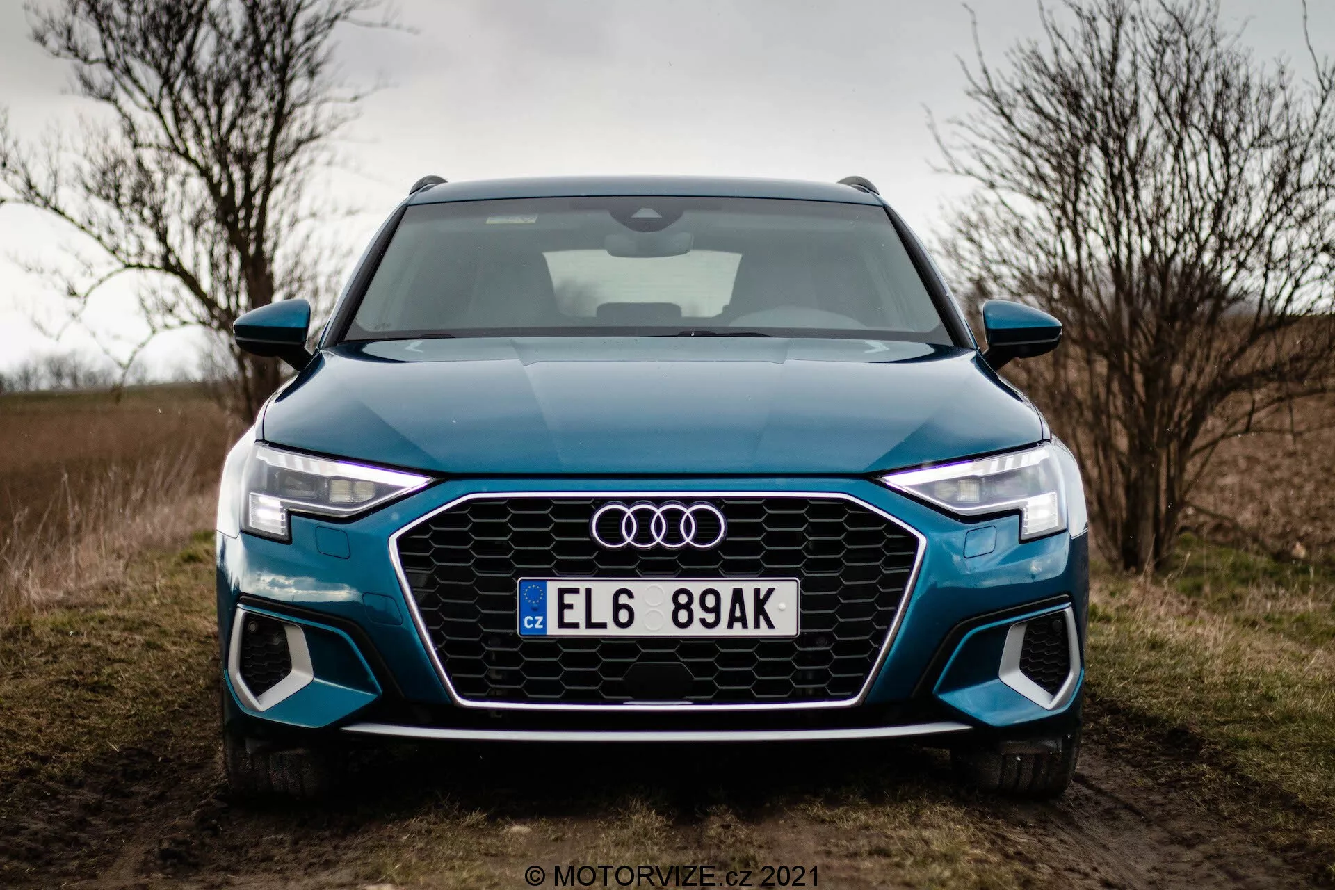 Vue de face d'une Audi A3 Sportback 2020 photographiée sous un angle bas. L'image se concentre sur l'avant du véhicule, avec la calandre Singleframe octogonale à motifs en nid d'abeille, les phares LED angulaires et nets avec des feux de jour distinctifs, et les grandes prises d'air intégrées dans le pare-chocs avant sculpté. Les quatre anneaux Audi sont montés au centre de la calandre, avec une plaque d'immatriculation de style européen en dessous. La voiture, d'un bleu vif, est garée au bord d'une route herbeuse avec un ciel couvert en arrière-plan, suggérant une saison automnale ou hivernale.