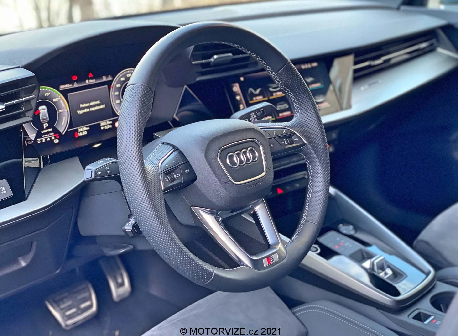 Vue intérieure d'une Audi A3 Sportback 2020 se concentrant sur le volant et la console centrale. Le volant sportif à trois branches, avec une partie inférieure plate, est doté de prises en cuir perforé, de boutons multifonctions, d'un emblème Audi et d'un badge 'S'. Derrière le volant, on aperçoit partiellement le cluster numérique, et à droite, l'écran tactile de l'Interface Multimédia Centrale (MMI) est partiellement visible. La console centrale comprend un levier de vitesse de transmission automatique, plusieurs boutons, un pavé tactile, deux porte-gobelets et de petits compartiments de rangement. Les matériaux intérieurs présentent des accents en métal brossé de qualité supérieure et des finitions de haute qualité, soulignant le luxe et la technologie avancée de la voiture.