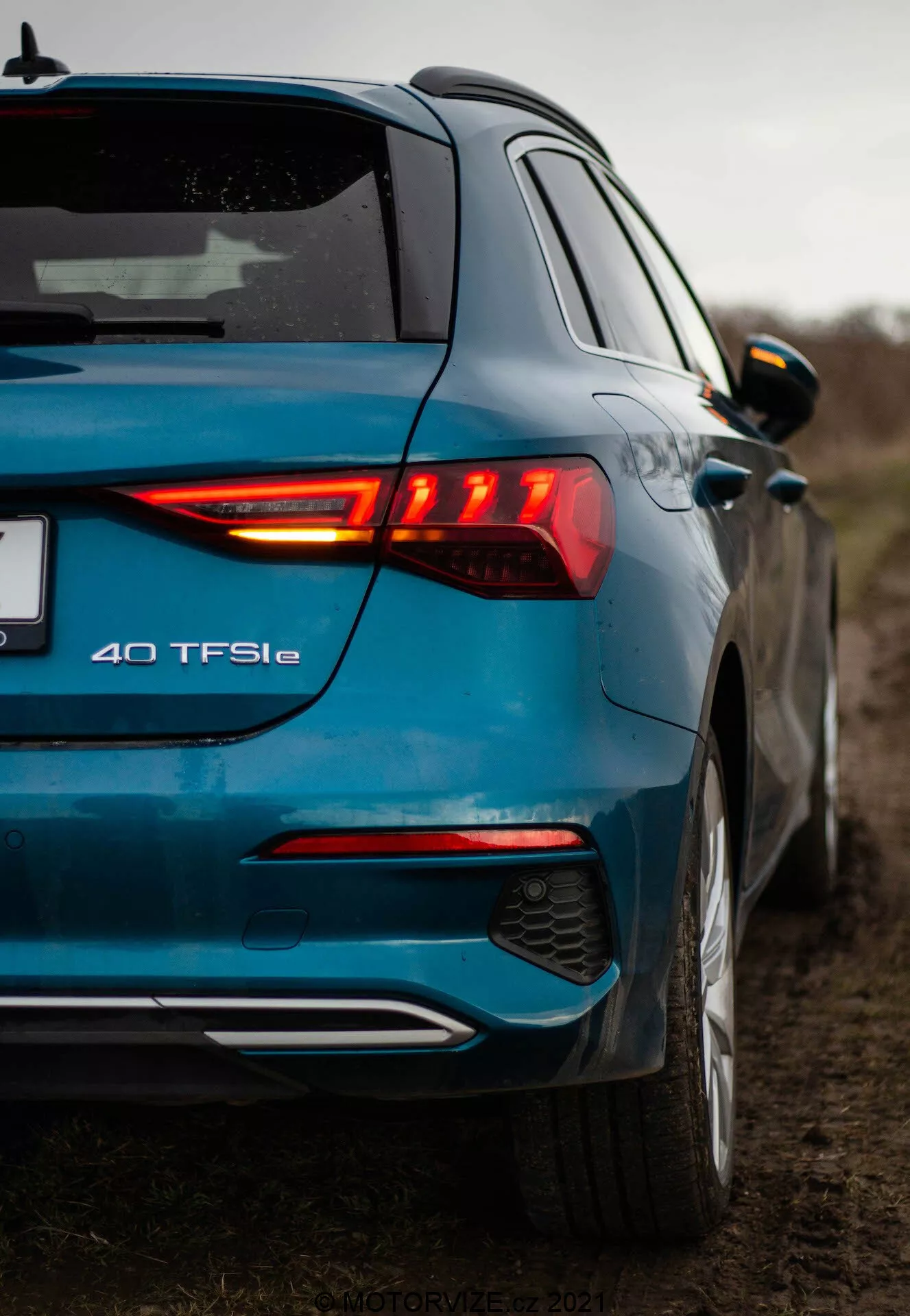 Une prise de vue en contre-plongée de l'Audi A3 2020 bleue moderne (A3 Sportback) prise d'une perspective arrière trois quarts, mettant en évidence le groupe de feux arrière à LED allumés, le badge du modèle 40 TFSIe, le pare-chocs arrière bicolore avec diffuseur intégré et capteurs de stationnement, la finition métallisée, le becquet de toit, les rétroviseurs latéraux assortis à la carrosserie avec clignotants intégrés et un soupçon de la porte avant et du profil latéral. La photographie met en valeur les éléments de design et les badges de la voiture dans un environnement rural avec un ciel couvert et une faible profondeur de champ.