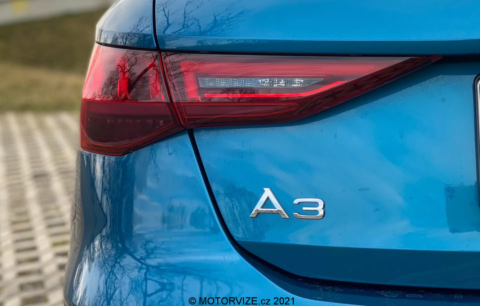 Cette image présente un gros plan, pris en angle bas, du coin arrière gauche d'une Audi A3 Sportback 2020 bleue. La photographie met en évidence le design élégant du feu arrière, qui a une orientation horizontale affûtée s'étendant sur le côté du véhicule, et la désignation du modèle 'A3' en chrome juste en dessous. Les éléments LED à l'intérieur du feu arrière mettent en lumière la technologie avancée d'éclairage de la voiture. La peinture brillante et la propreté générale du modèle montrent un entretien de haute qualité, avec un arrière-plan légèrement flou suggérant un cadre extérieur. Le filigrane '© MOTORVIZE.cz 2021' indique la propriété et l'origine de l'image.