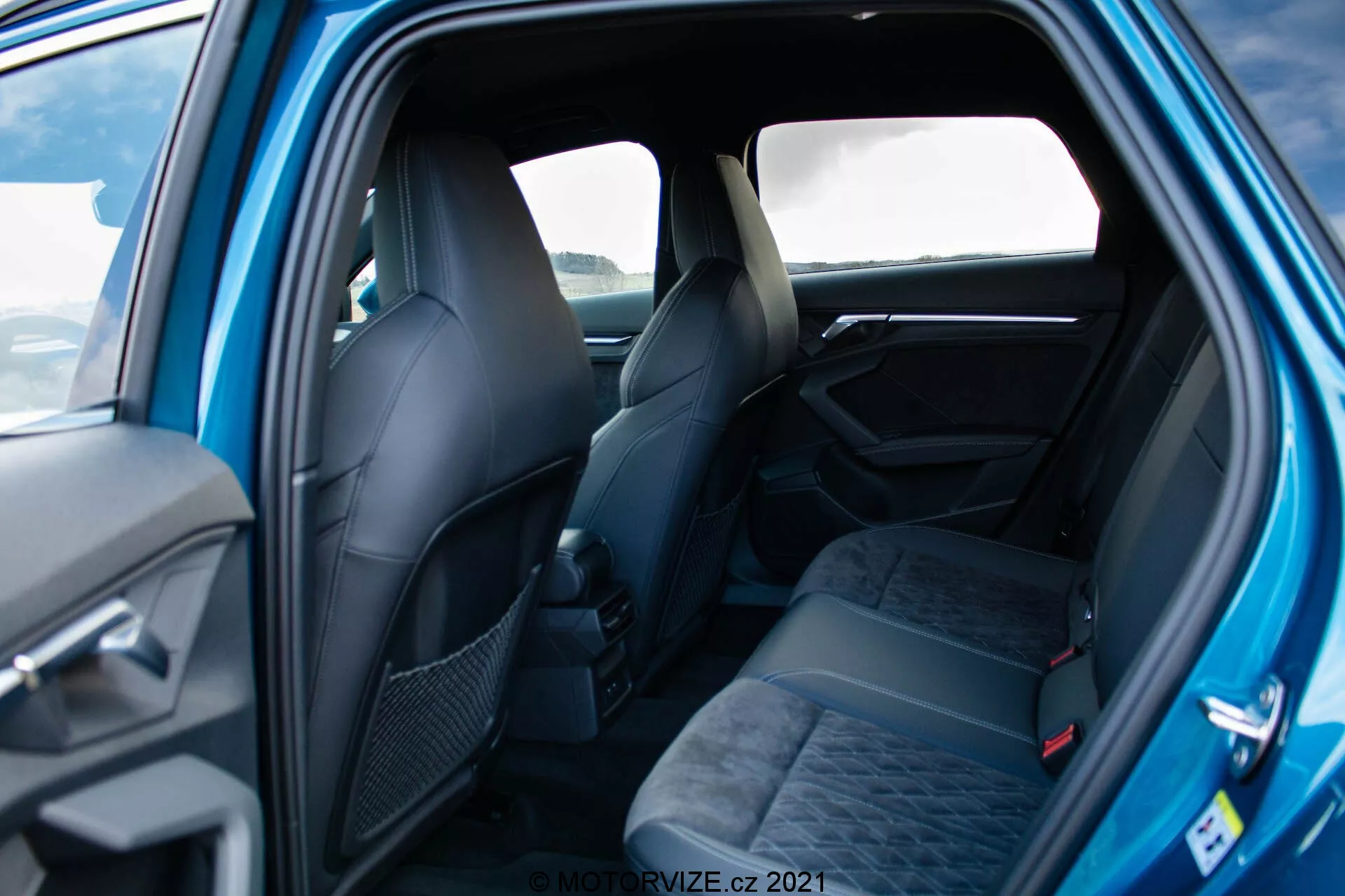 Photo de l'intérieur d'une Audi A3 Sportback 2020 vue du côté passager arrière, montrant un espace assis pour trois avec appuis-têtes et ceintures de sécurité, tous en tissu sombre à motifs. Les sièges avant, à demi visibles, affichent également un design ergonomique. Les éléments du panneau de porte visibles comprennent une poignée et des contrôles possibles. L'image bien éclairée met en avant une atmosphère élégante et premium avec des matériaux et des détails de conception de qualité.