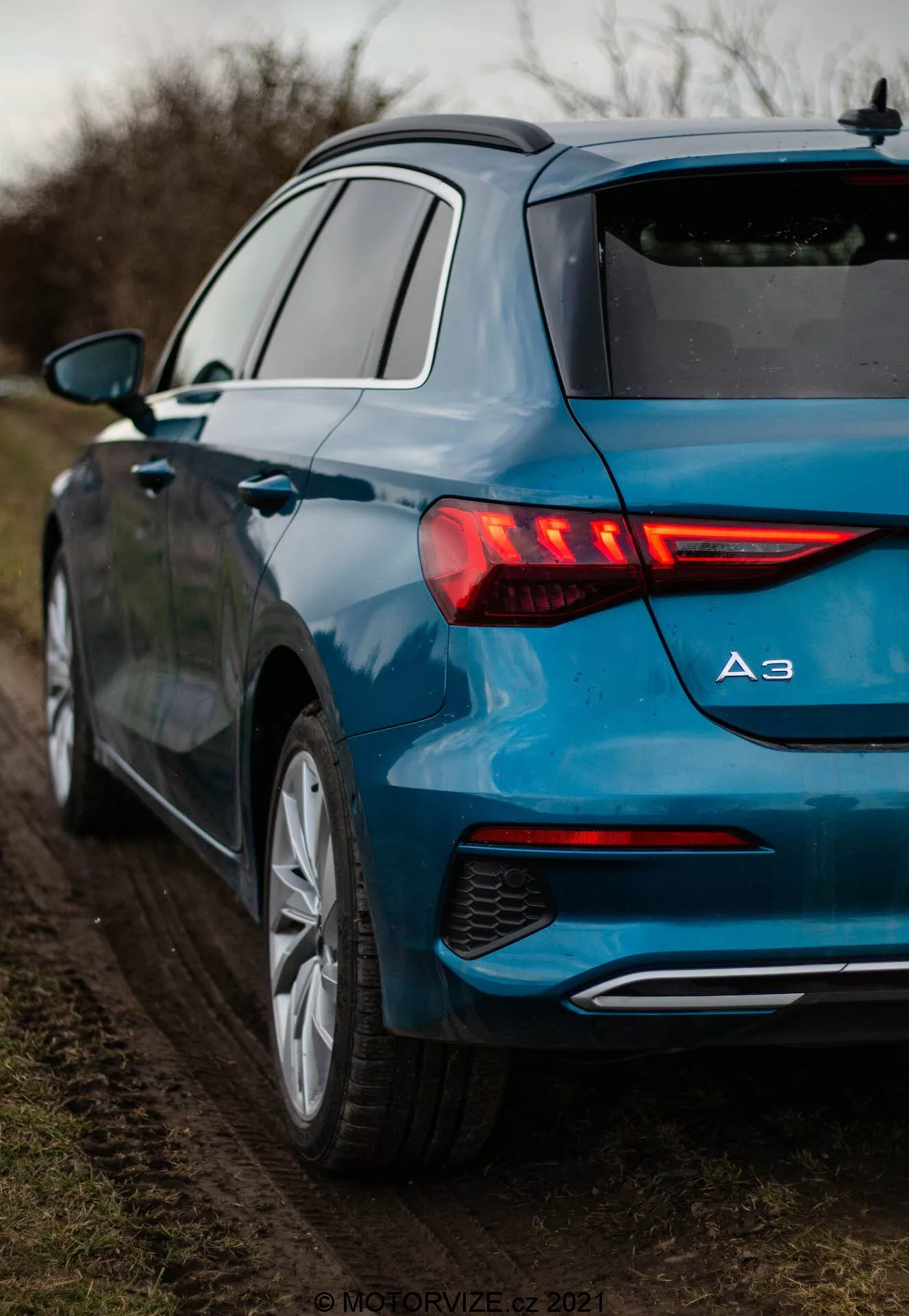 Vue détaillée d'une Audi A3 2020 Sportback bleu métallisé sous un angle arrière trois quarts, mettant en avant ses feux arrière à LED illuminés avec clignotants dynamiques, le badge du modèle affichant 'A3', des jantes en alliage à multiples rayons et une sortie d'échappement unique. La voiture est garée sur le bord d'une route non pavée entourée de végétation, sous un éclairage doux et diffus.