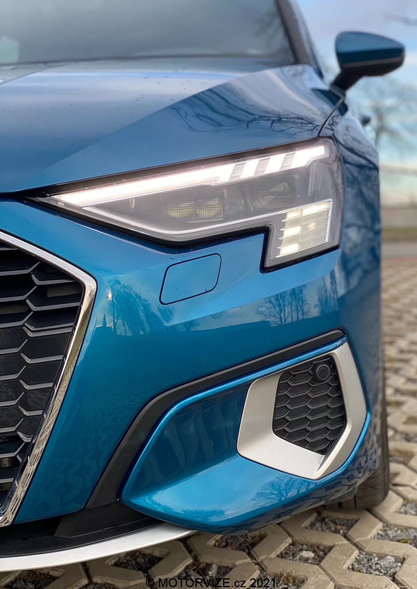 Gros plan sur le quart avant gauche d'une Audi A3 2020 bleue (A3 Sportback), mettant en valeur le phare LED angulaire élégant avec feux de jour, la calandre hexagonale en nid d'abeille proéminente et la prise d'air avec bord métallique. La prise de vue en contre-plongée souligne les éléments de design sportif, le design géométrique complexe des antibrouillards et le fond flou pour un focus sur les détails.