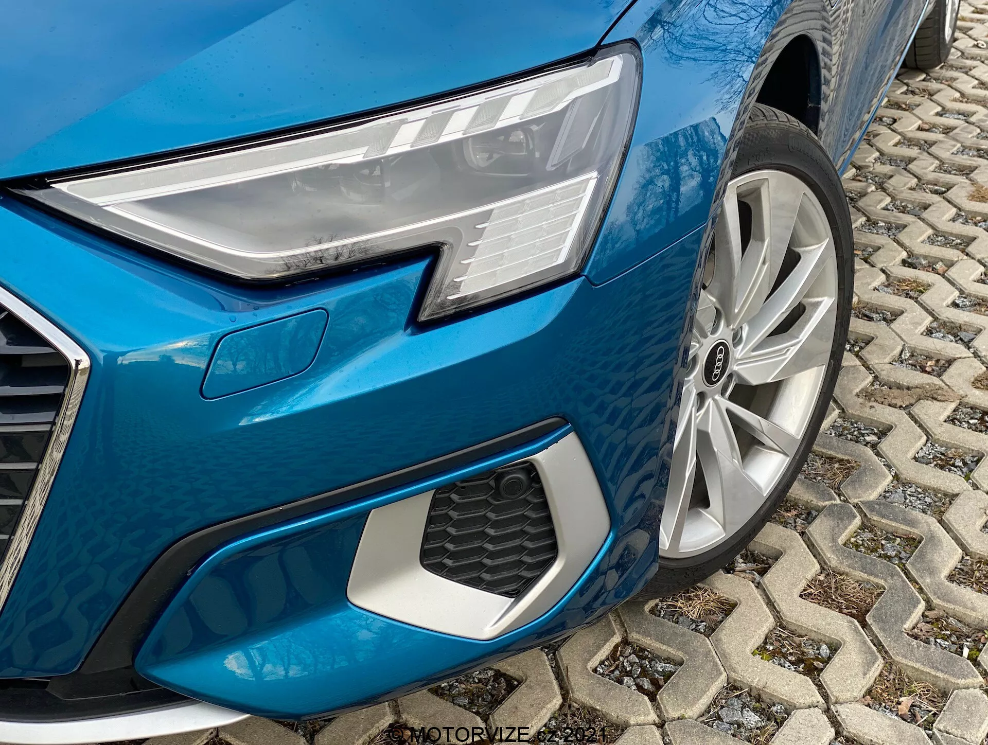 Vue en gros plan du quart avant gauche de l'Audi A3 2020 Sportback bleu métallique. L'image, prise sous un angle bas, met en évidence des éléments de conception clés, notamment le phare LED moderne avec des éléments lumineux individuels, la roue en alliage à rayons multiples, la face avant angulaire avec une grille en nid d'abeille proéminente et le pavage en pierre dans un environnement urbain.