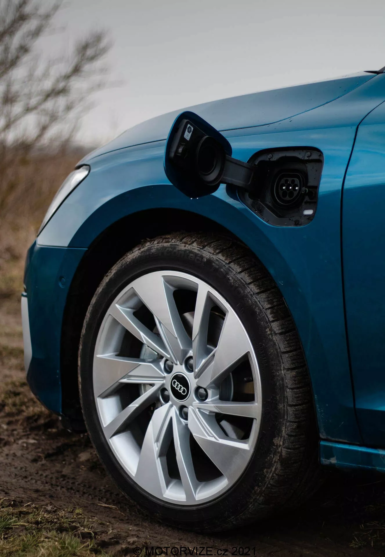 Gros plan du quart avant côté conducteur d'une Audi A3 2020 Sportback bleue, mettant en évidence la roue et le port de charge. L'image, prise d'un angle bas vers l'avant, montre le port de charge ouvert au-dessus de la roue avant, avec un câble de recharge connecté. La faible profondeur de champ se concentre nettement sur la roue et le port de charge, révélant le logo distinctif à anneaux entrelacés d'Audi et la conception de roue à plusieurs rayons avec un pneu à profil bas, le tout sous un éclairage légèrement atténué, potentiellement au crépuscule.