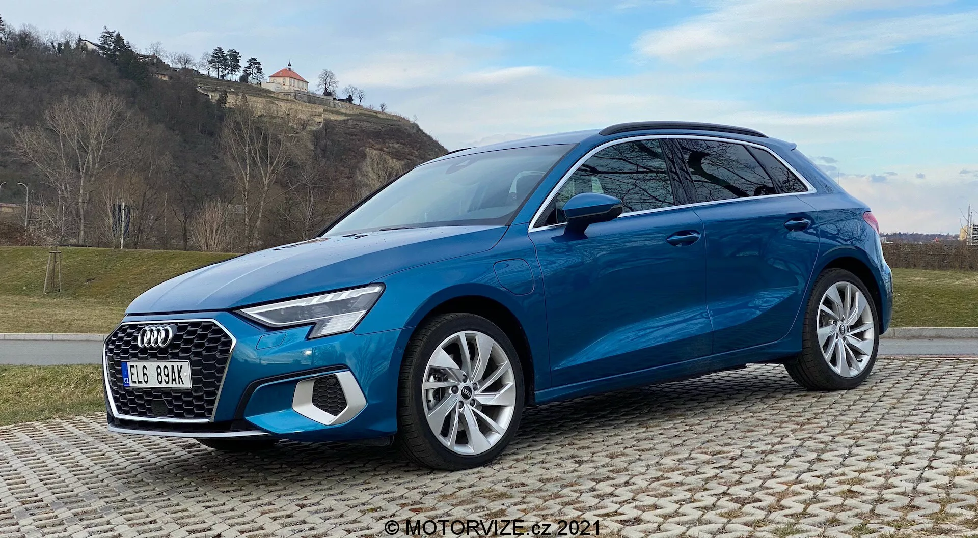 Vue de trois quarts avant d'une Audi A3 Sportback 2020 bleu vif sur des pavés, montrant des éléments de design distinctifs tels que des phares à LED, une calandre proéminente, des jantes en alliage à plusieurs branches et des systèmes de sécurité avancés. Capturé avec un arrière-plan herbeux et vallonné et un ciel partiellement nuageux.