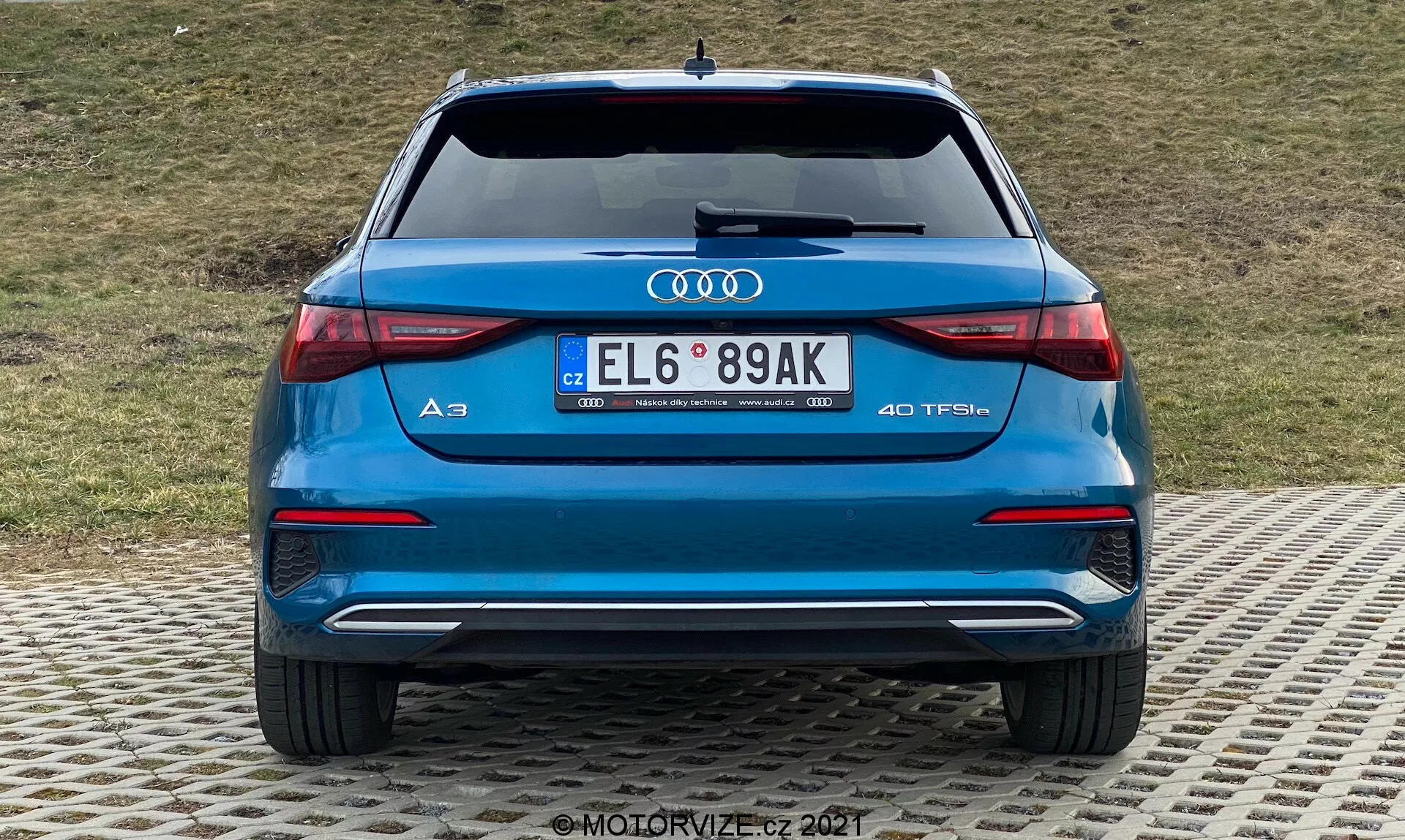 Vue arrière d'une Audi A3 2020 Sportback bleue, présentant le pare-chocs arrière, le coffre, les feux arrière, les tuyaux d'échappement et la lunette arrière. Logo Audi bien visible et désignation du modèle 'A3' avec le badge '40 TFSI' indiquant un moteur turbo essence. Feux arrière LED avec un design horizontal élégant, doubles tuyaux d'échappement, pare-chocs arrière avec élément de diffuseur et capteurs de stationnement, et plaque d'immatriculation de style européen. Arrière-plan avec une pente herbeuse.