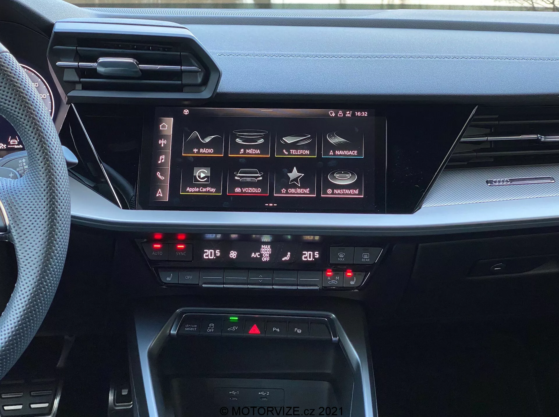 Vue tableau de bord intérieur de l’Audi A3 Sportback 2020, mettant en avant son système d'infodivertissement avancé et ses commandes de climatisation. L'image capture un grand écran tactile horizontal avec des fonctionnalités telles que la navigation, les médias, la connectivité téléphonique et les réglages de la voiture, indiquant des capacités d'infodivertissement complètes, dont Apple CarPlay et Android Auto. Sous l'écran, les commandes numériques de la climatisation bi-zone indiquent des réglages à 20 degrés Celsius pour le conducteur et le passager. Le tableau de bord comprend également un espace de recharge sans fil et des fentes d'aération minces et horizontales, illustrant le design moderne des intérieurs automobiles. La photo est marquée du filigrane 'MOTORVIZE.CZ 2021'.