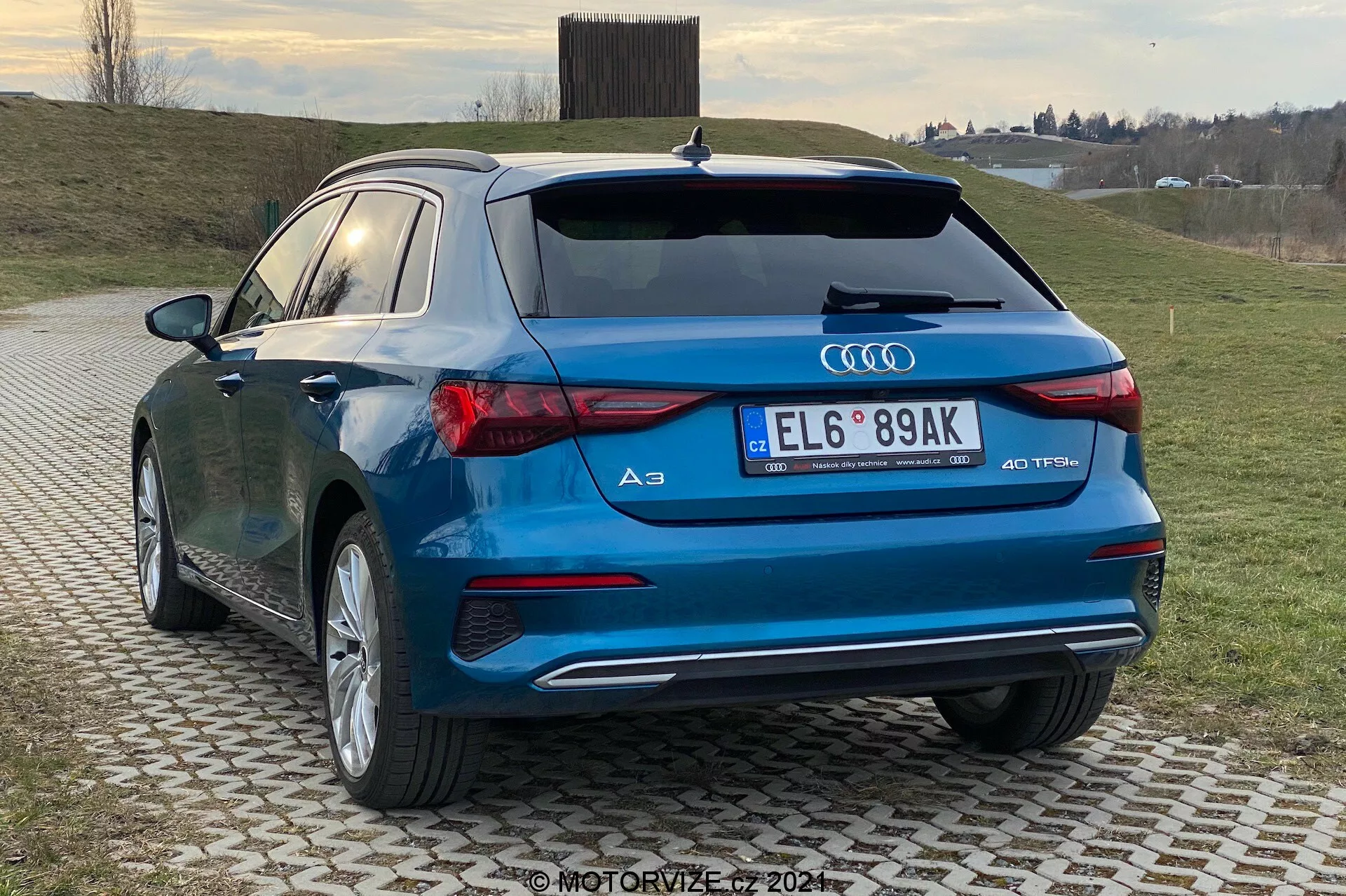 L'image capture une berline compacte moderne, l'Audi A3 Sportback 2020, d'une vue arrière trois quarts, mettant en valeur le profil arrière et latéral du véhicule. Des éléments de design distinctifs tels que les feux arrière à LED, les sorties d'échappement doubles, l'emblème des anneaux Audi et le badge '40 TFSI' sont visibles, indiquant une variante sportive S-line. La voiture, d'une finition bleue brillante, est garée sur un pavé à motifs dans un cadre rural ou semi-rural avec un fond herbeux et un ciel couvert.