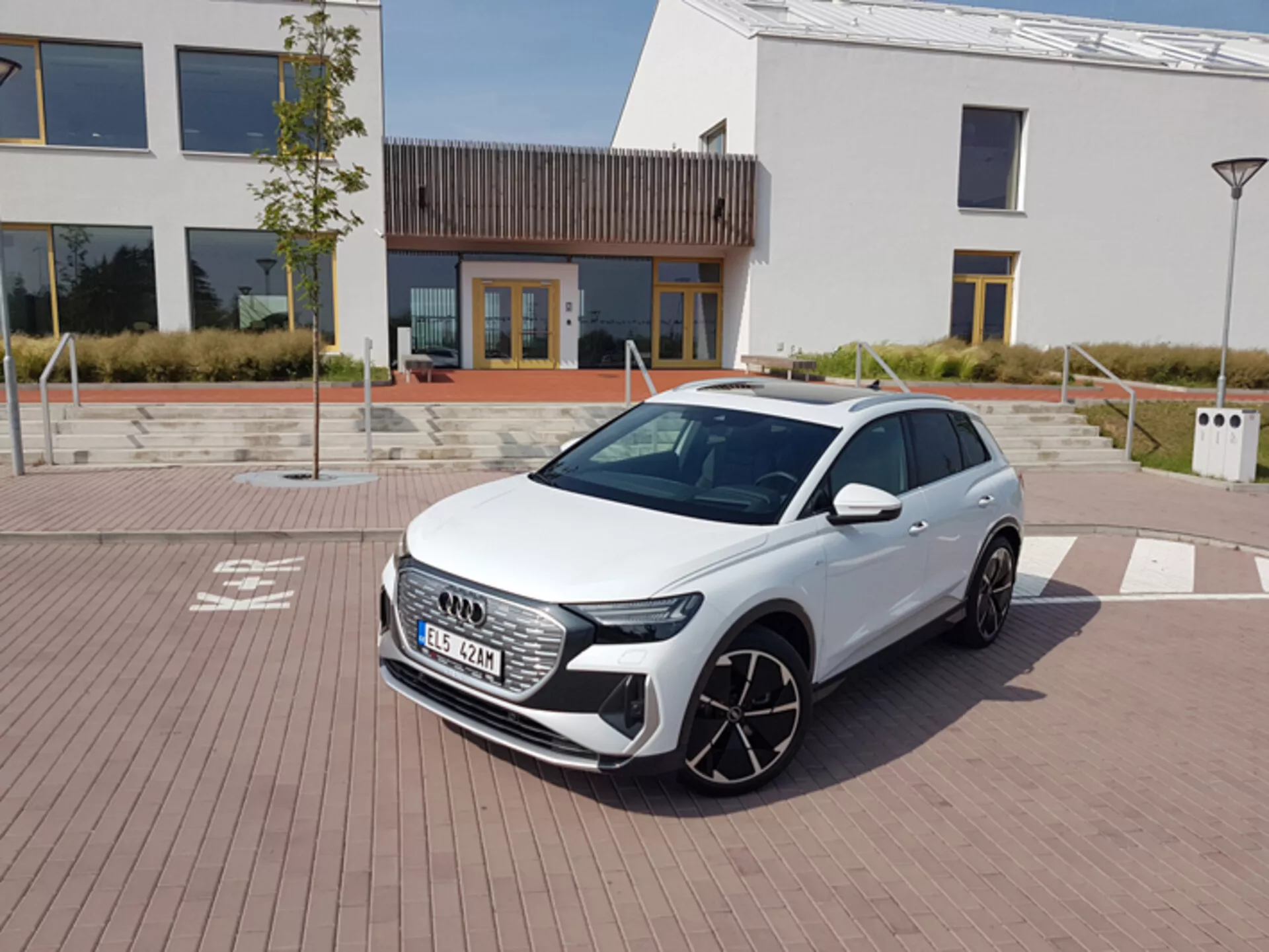 Audi Q4 e-tron 2021 en blanc, présentant ses phares LED modernes, sa calandre frontale hexagonale et ses grandes roues en alliage, garée sur un pavage à motif de briques avec une architecture urbaine et une station de charge en arrière-plan, sous un ciel clair à midi.