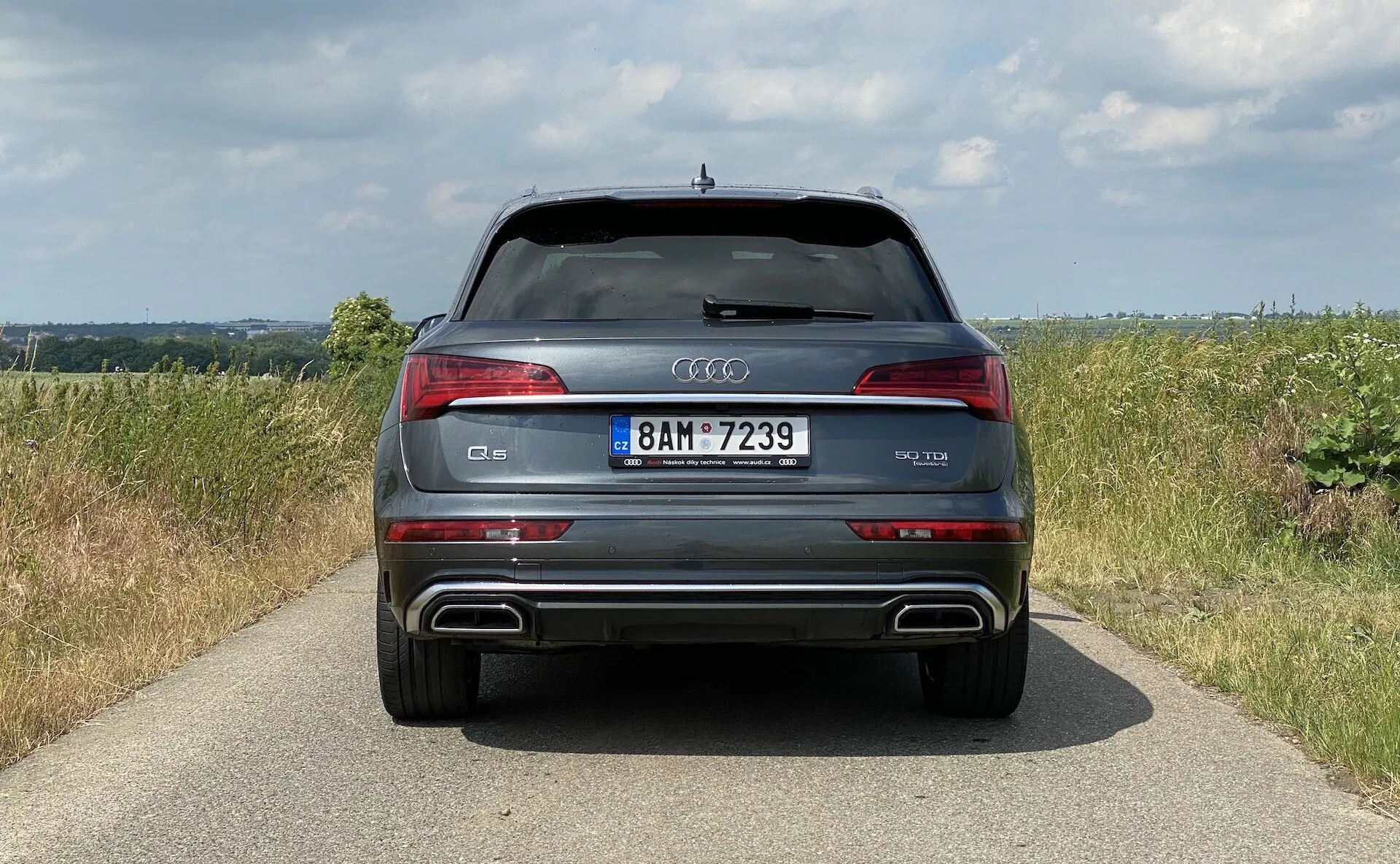 Vue arrière d'un SUV moderne Audi Q5 2020 Sportback mettant en avant la symétrie de son design avec des feux arrière à LED distinctifs, pare-chocs arrière avec accents métalliques et ports d'échappement doubles intégrés, et une bande chromée au-dessus de la plaque d'immatriculation. Positionné sur un pavé étroit au milieu d'un cadre campagnard avec des champs et un ciel couvert, montrant ses caractéristiques de luxe et sportives, y compris les capteurs de parking et un aileron de toit.