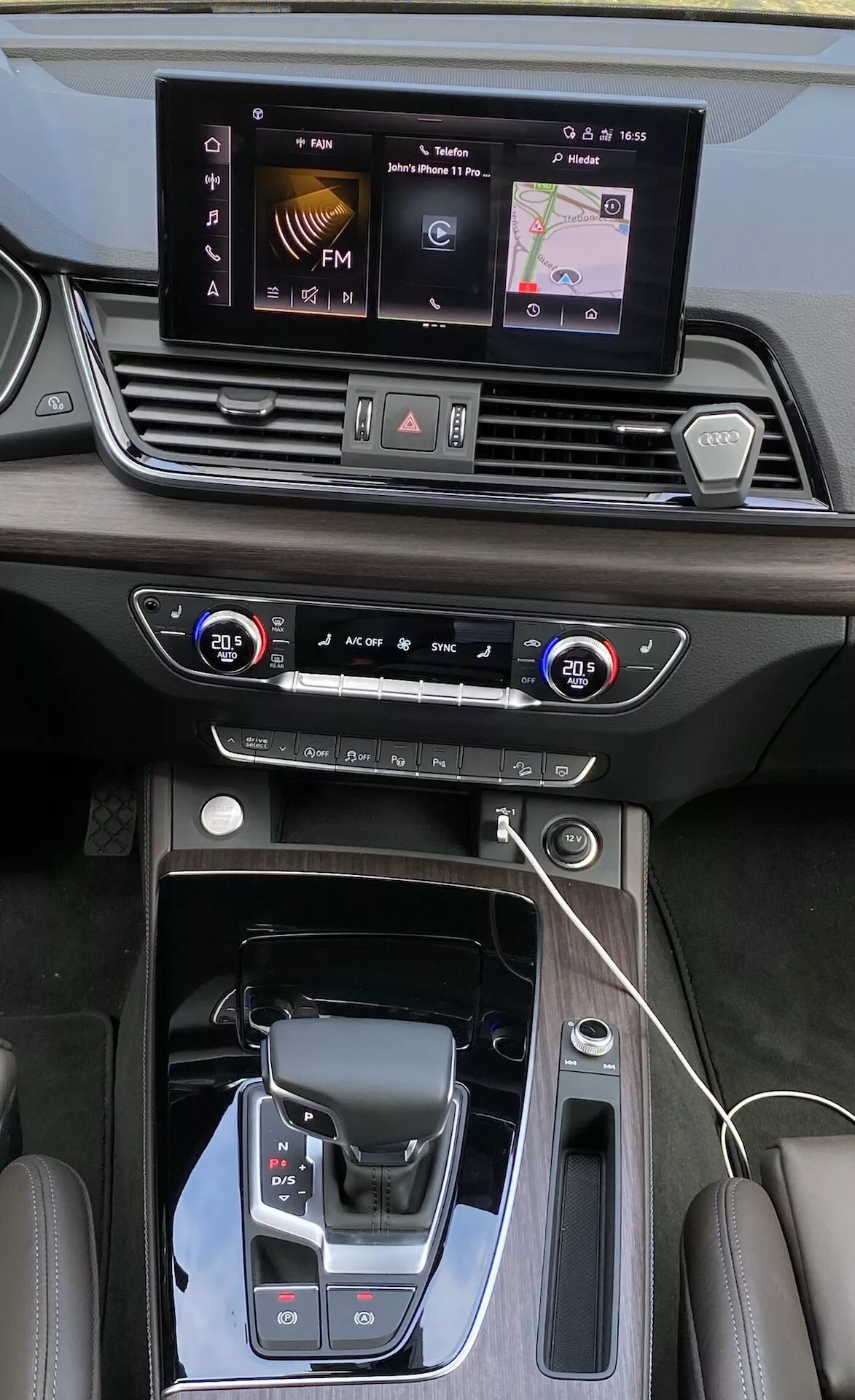 Vue en plongée de la console centrale et du tableau de bord de l'Audi Q5 2020 Sportback, présentant des caractéristiques détaillées telles que l'écran tactile d'infodivertissement, les commandes de climatisation à deux zones, le sélecteur de vitesse moderne avec un soufflet en cuir et des coutures en cuir luxueuses. La perspective met en évidence le design élégant, les accents en noir piano et métal et les technologies avancées, y compris un chargeur sans fil et la sélection des modes de conduite.