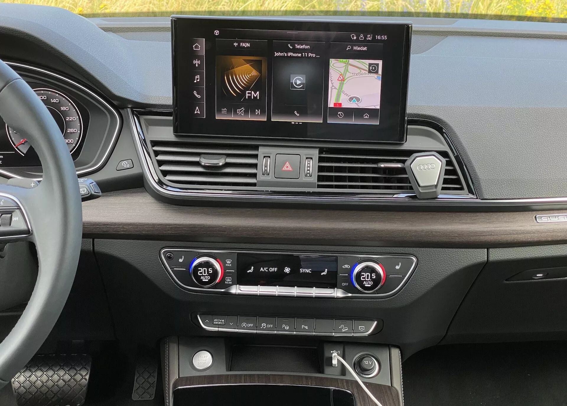 Vue du tableau de bord intérieur de l'Audi Q5 2020 Sportback depuis une perspective à hauteur d'œil. L'image met en avant l'écran large de l'interface multimédia avec des options comme la radio FM, la navigation et les réglages du véhicule. En dessous, on peut voir quatre bouches d'aération disposées symétriquement, une configuration centrale de boutons et de cadrans pour les feux de détresse, le dégivrage et les réglages d'accès rapide. Le système de contrôle climatique à double zone avec affichages numériques de la température est réglé à 20,5 degrés Celsius pour le conducteur et le passager. La section inférieure révèle un espace de rangement avec des ports USB possibles, une entrée auxiliaire et un bouton de démarrage/arrêt pour la technologie d'allumage sans clé. Les matériaux et le design de luxe comprennent des finitions en bois et des accents métalliques polis.