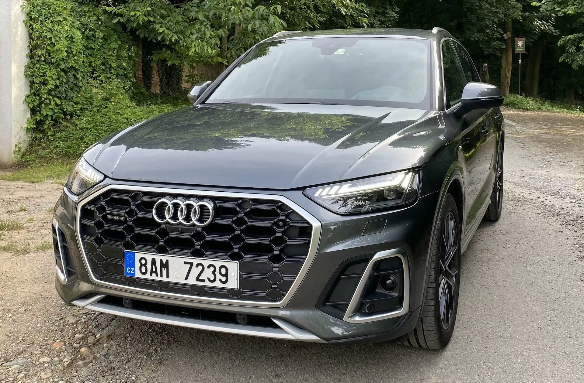 Vue avant trois quarts d'un Audi Q5 Sportback 2020 montrant le côté avant gauche avec la grille hexagonale à alvéoles caractéristique, le logo Audi, les phares LED angulaires et élégants, et les grandes prises d'air sur le pare-chocs inférieur. La voiture est garée sur une route entourée d'arbres, reflétant sa peinture sombre et brillante. Le capot sculpté avec des lignes aérodynamiques vers l'emblème et la roue avant en alliage à plusieurs rayons soulignent son design sportif et moderne.
