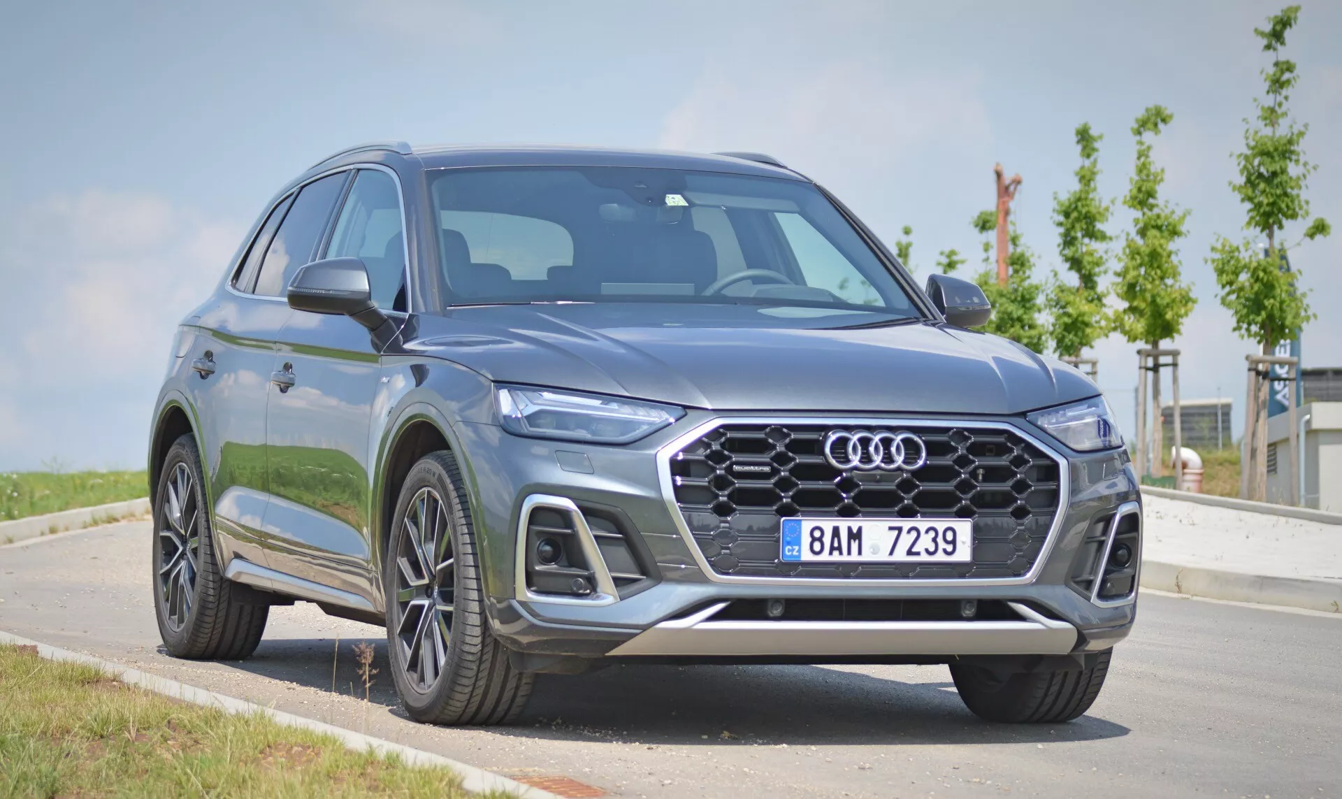 L'image présente un Audi Q5 Sportback 2020, un SUV crossover de luxe moderne. Capturé d'un angle frontal bas à trois quarts, il met en avant la grille hexagonale proéminente, les phares LED avec des feux de jour distinctifs, les roues en alliage et les lignes sculptées de la carrosserie. Le véhicule est garé sur une surface en asphalte avec un ciel dégagé et de la verdure en arrière-plan, offrant une vue détaillée de sa finition métallique grise ou bleue et de ses caractéristiques de design haut de gamme.