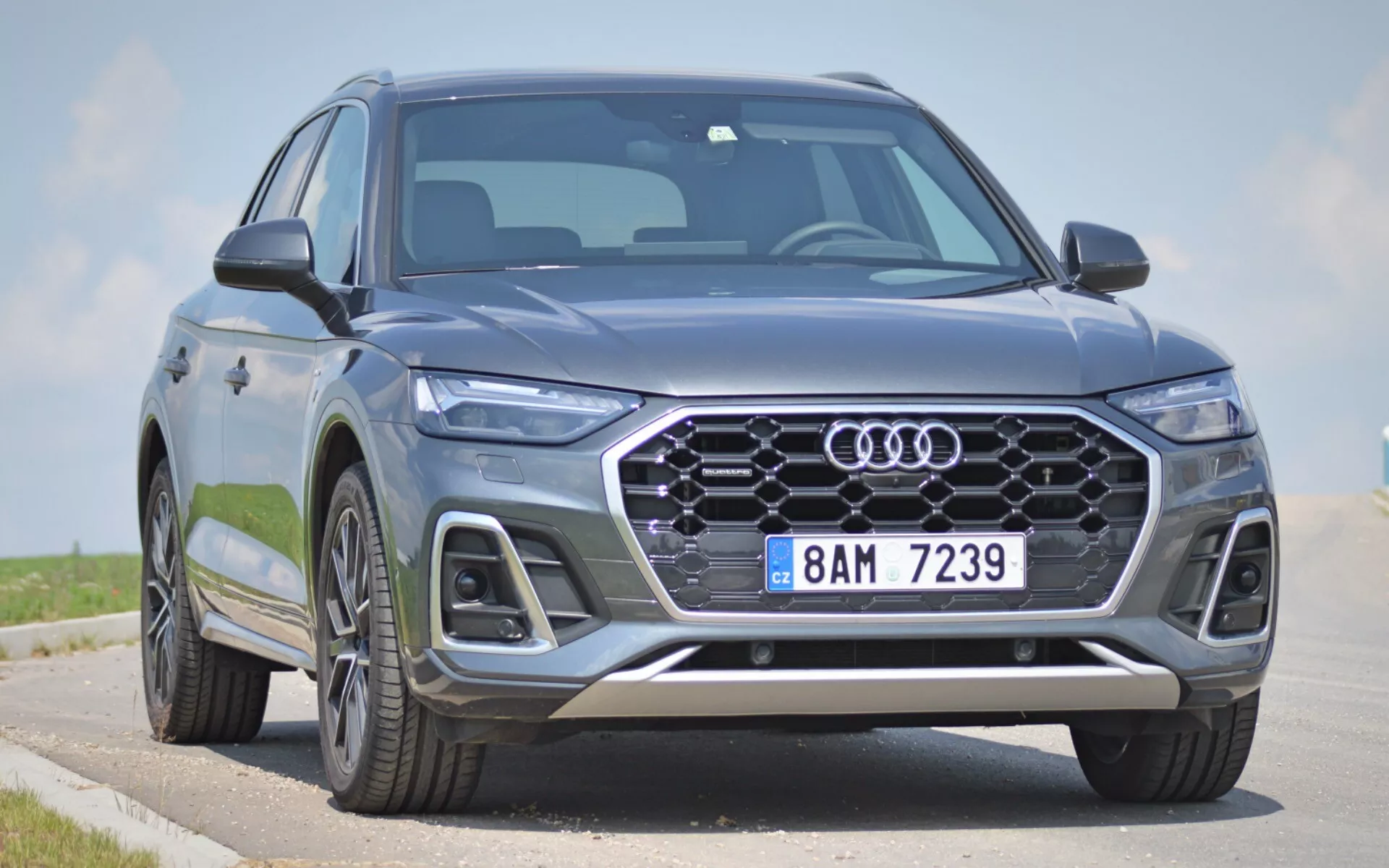 Audi Q5 2020 moderne (Q5 Sportback) SUV capturé sous un angle de trois quarts avant bas, avec la face avant, la calandre hexagonale avec emblème à quatre anneaux, des phares LED élégants et une partie du profil latéral. La grande calandre avec un design à motifs, les phares antibrouillard intégrés, les prises d'air et les éléments en argent sur le pare-chocs avant ajoutent un attrait esthétique et fonctionnel. Les technologies avancées d'éclairage et d'assistance à la conduite sont suggérées par le design pointu des phares et la configuration de la calandre. Les grandes roues en alliage et les lignes dynamiques de la carrosserie indiquent une présence premium et robuste. Stationné sur une route avec un ciel dégagé en arrière-plan, les caractéristiques frontales et les éléments stylistiques agressifs du véhicule sont mis en avant.