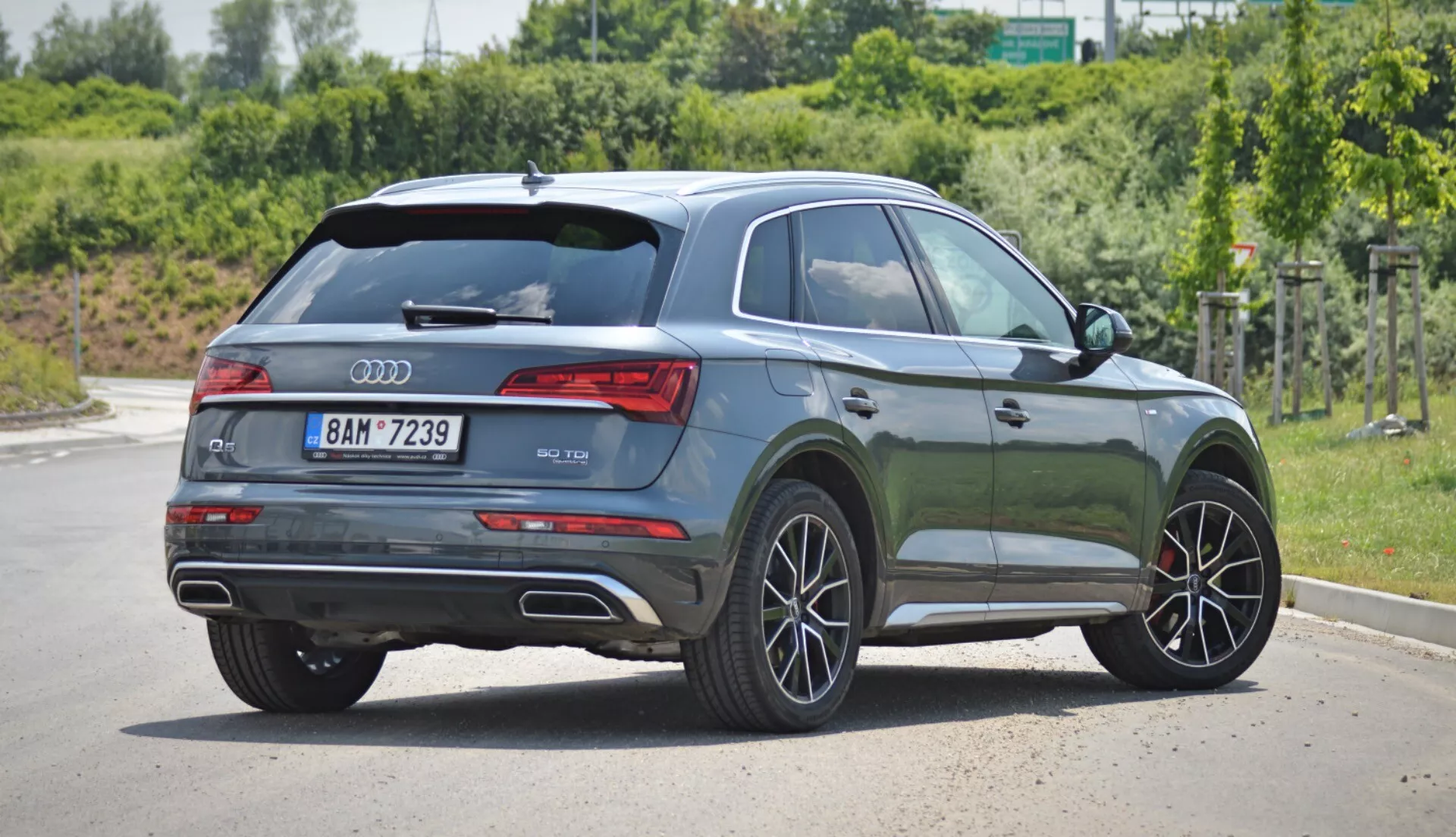 L'image montre un SUV moderne Audi Q5 2020 Sportback garé sur une surface pavée à la lumière du jour, vu sous un angle arrière trois quarts. La porte arrière arbore le logo Audi et la désignation du modèle, avec des feux arrière horizontaux élégants s'intégrant dans le profil latéral. La peinture métallique brillante vert foncé reflète la lumière, mettant en valeur ses contours. Les vitres latérales du passager sont teintées, suggérant une catégorie premium. La ligne de toit s'incline doucement vers l'arrière, donnant une apparence sportive, accentuée par un aileron de toit. Les roues en alliage avec un design multi-branches et les freins à disque visibles suggèrent une finition améliorée. La garde au sol du SUV et les protections de carrosserie le long des seuils latéraux et autour des passages de roues indiquent sa polyvalence et sa capacité tout-terrain. Les rails de toit argentés suggèrent une fonctionnalité supplémentaire tandis que les poignées de porte et les rétroviseurs latéraux sont de la même couleur que la carrosserie. Les sorties d'échappement doubles dans le pare-chocs arrière suggèrent un moteur puissant.