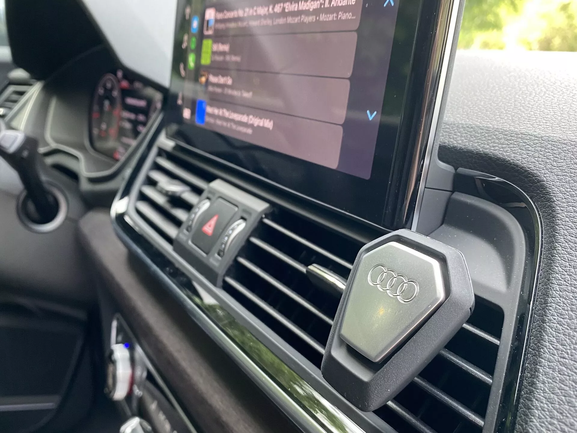 Vue rapprochée depuis le côté du conducteur montrant le tableau de bord central d'une Audi Q5 Sportback 2020. L'accent est mis sur les aérateurs centraux et l'écran d'infodivertissement affichant une lecture multimédia avec une liste de fichiers audio, soulignant les capacités multimédia du véhicule. Les aérateurs sont rectangulaires avec une finition métallique et comportent des boutons avec le logo Audi. Le tableau de bord présente une surface texturée de qualité premium, en ligne avec le positionnement de luxe de la marque Audi, et il n'y a pas de commande manuelle de climatisation visible, ce qui suggère un système de contrôle climatique automatique.