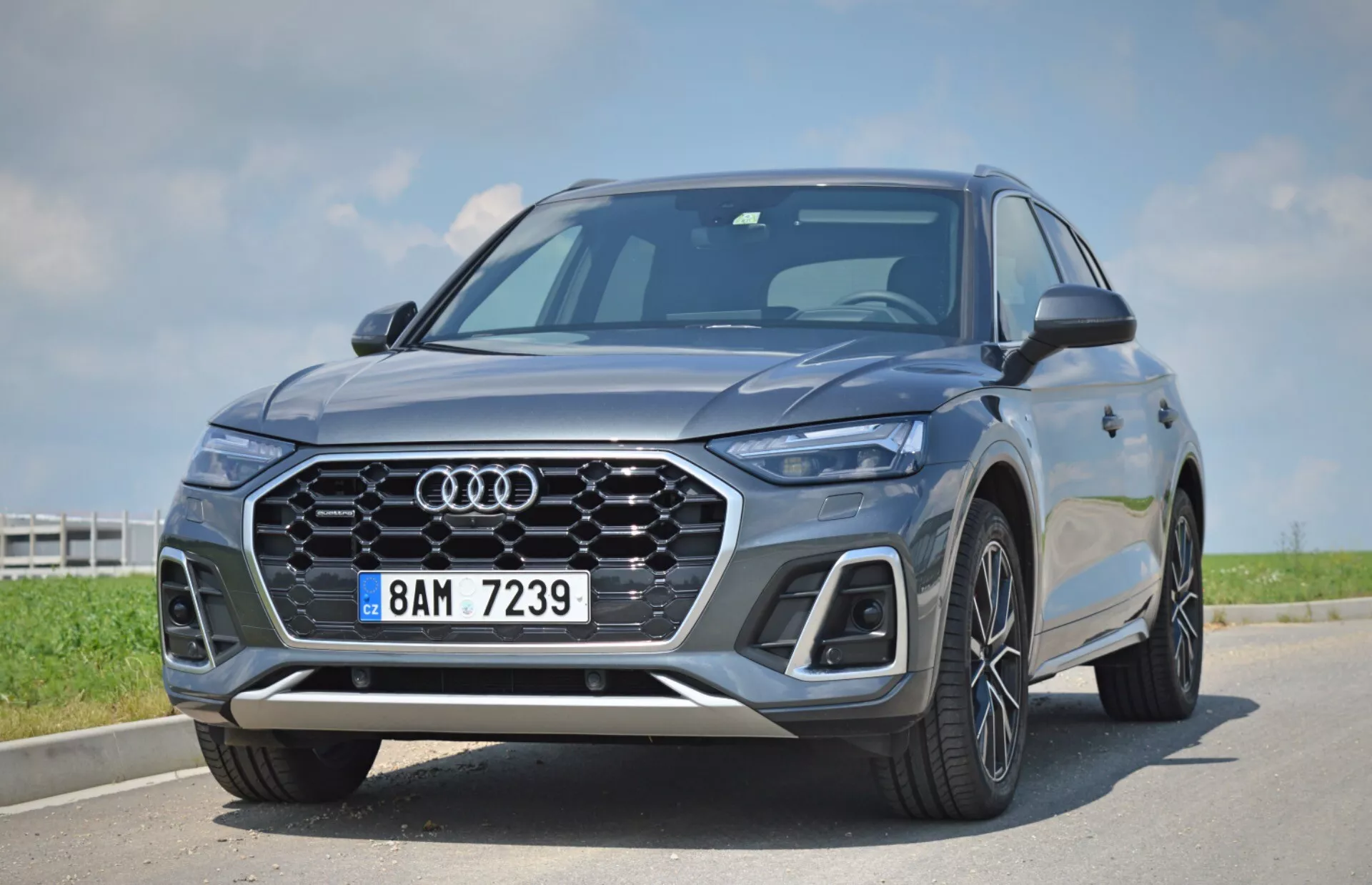 L'image présente un SUV moderne Audi Q5 Sportback 2020 capturé sous un angle bas et rapproché, mettant en valeur l'avant du véhicule et la calandre. Les caractéristiques distinctives incluent l'emblème iconique des quatre anneaux d'Audi, une calandre à cadre unique avec un design en nid d'abeille, des phares à LED élégants avec des feux de jour distinctifs, des capteurs intégrés dans le pare-chocs avant indiquant des systèmes avancés d'aide à la conduite, de grandes roues en alliage multi-brins, des lignes de carrosserie nettes et des rétroviseurs latéraux aérodynamiques. Le véhicule est garé sous un ciel dégagé, mettant en évidence son design luxueux et technologiquement avancé.