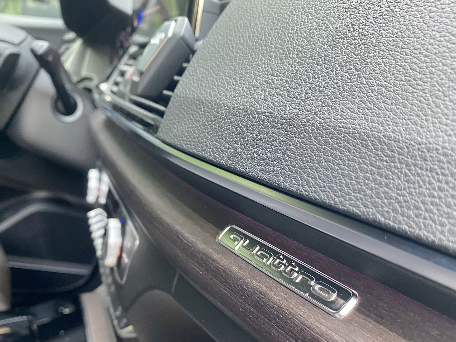 Vue en gros plan de l'intérieur de l'Audi Q5 2020 Sportback, concentrée sur l'emblème 'QUATTRO' à hauteur des yeux, mettant en valeur le système à quatre roues motrices d'Audi. Doté d'une finition brillante sur l'emblème et de matériaux texturés contrastés, l'image est prise sous un angle oblique avec une faible profondeur de champ, montrant des détails du tableau de bord et du panneau de commande central dans un arrière-plan flou.