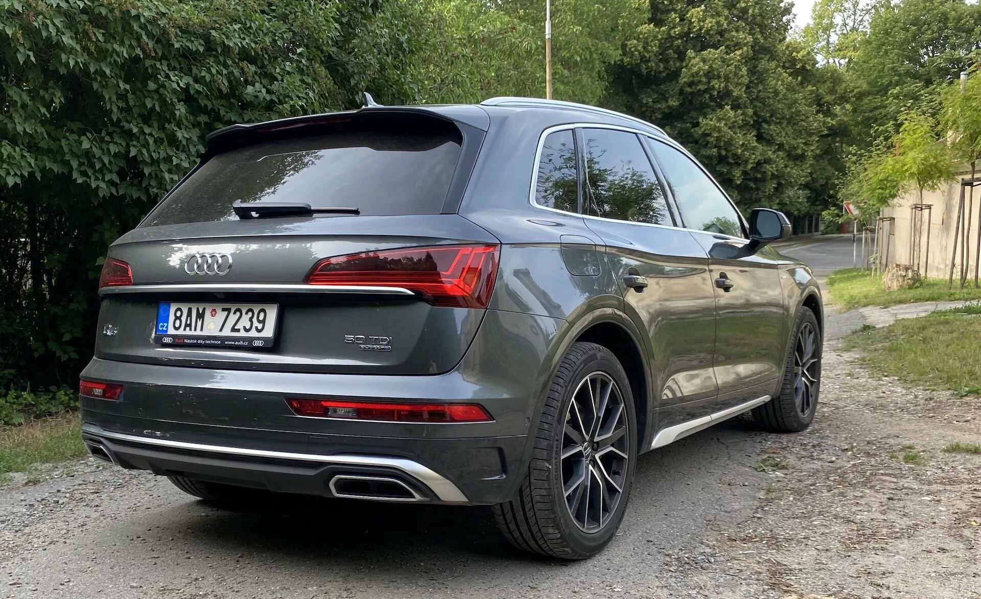 Vue arrière en angle d'un SUV Audi Q5 Sportback 2020 gris garé sur une surface en gravier à côté de végétation verte. L'image se concentre sur le profil arrière et latéral de la voiture, montrant des feux arrière LED modernes, des doubles sorties d'échappement, le logo Audi en anneaux, les badges du modèle 'Q7' et de la variante '45 TDI', des jantes en alliage à plusieurs rayons, des vitres teintées, un aileron de toit et des capteurs de stationnement ou une caméra potentiellement présentes. L'éclairage naturel avec des ombres douces suggère un lever ou un coucher de soleil.
