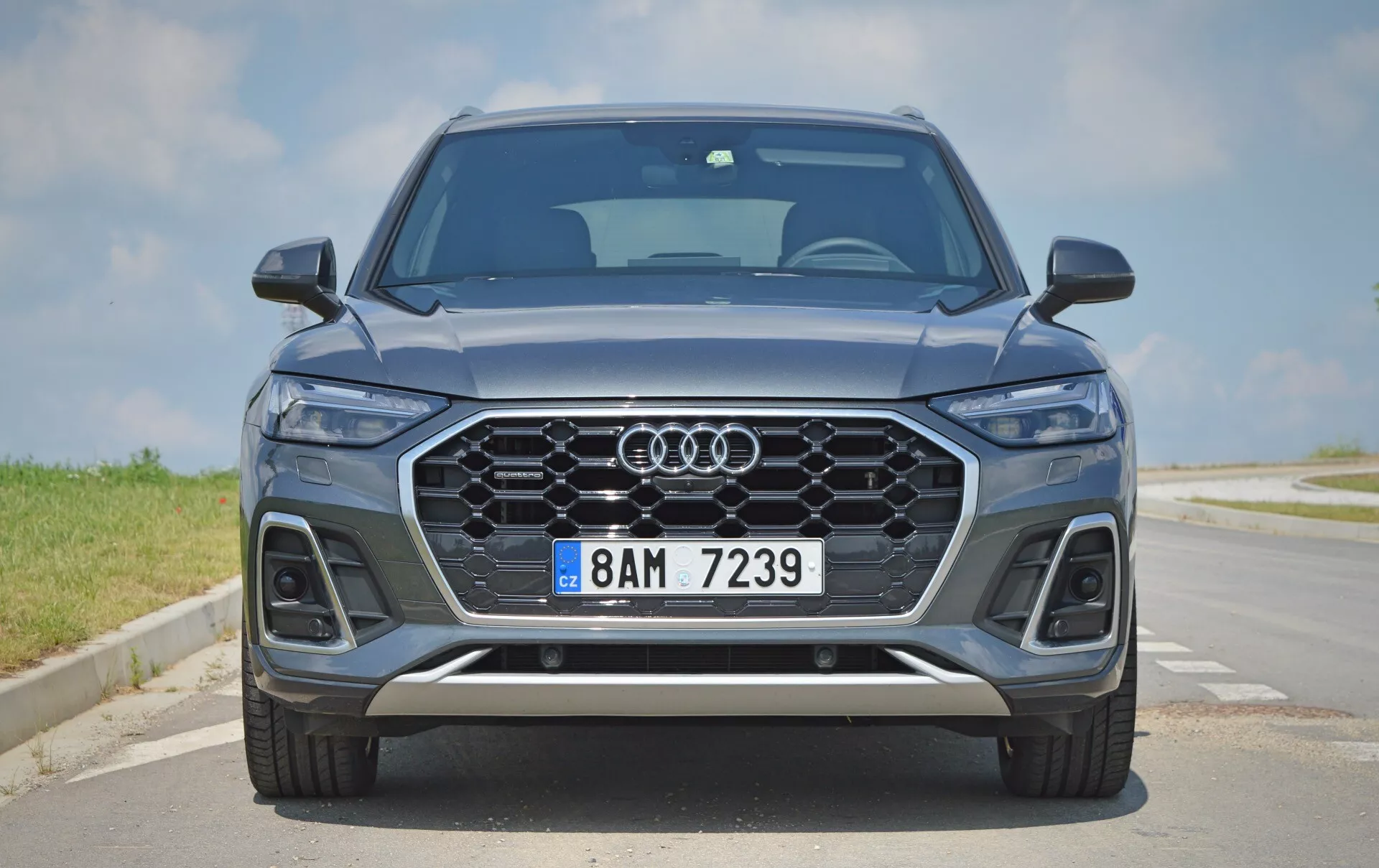 Vue frontale d'un moderne SUV Audi Q5 2020 Sportback mettant en valeur son avant élégant et symétrique, avec la célèbre grille Audi, des phares à LED, des capteurs de stationnement, une peinture bleu métallique, des jantes en alliage et des détails en finition argentée. La scène se déroule sur une route pavée sous un ciel clair.