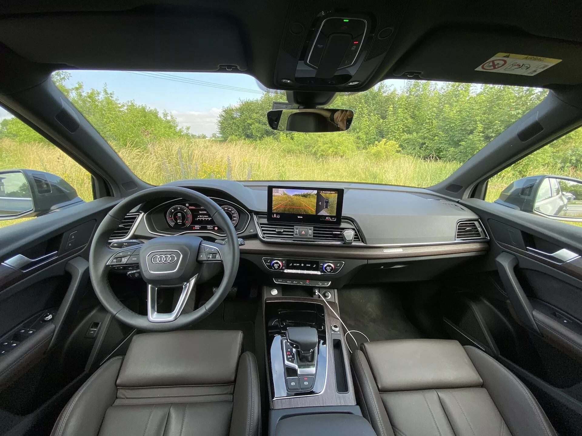 Intérieur de l'Audi Q5 Sportback 2020 photographié depuis la perspective du siège du conducteur, montrant un tableau de bord luxueux et technologiquement avancé. L'image met en avant des sièges en cuir noir, une console centrale avec un accoudoir en cuir, un levier de vitesses automatique entouré de boutons de commande, un écran tactile horizontal d'infotainment avec navigation GPS, un contrôle climatique à double zone, un écran numérique pour le conducteur derrière un volant au logo Audi avec commandes intégrées. Le design présente un minimalisme avec des accents en métal brossé, offrant une visibilité complète à travers la vitre et le pilier de la voiture, indiquant qu'elle est garée en extérieur.