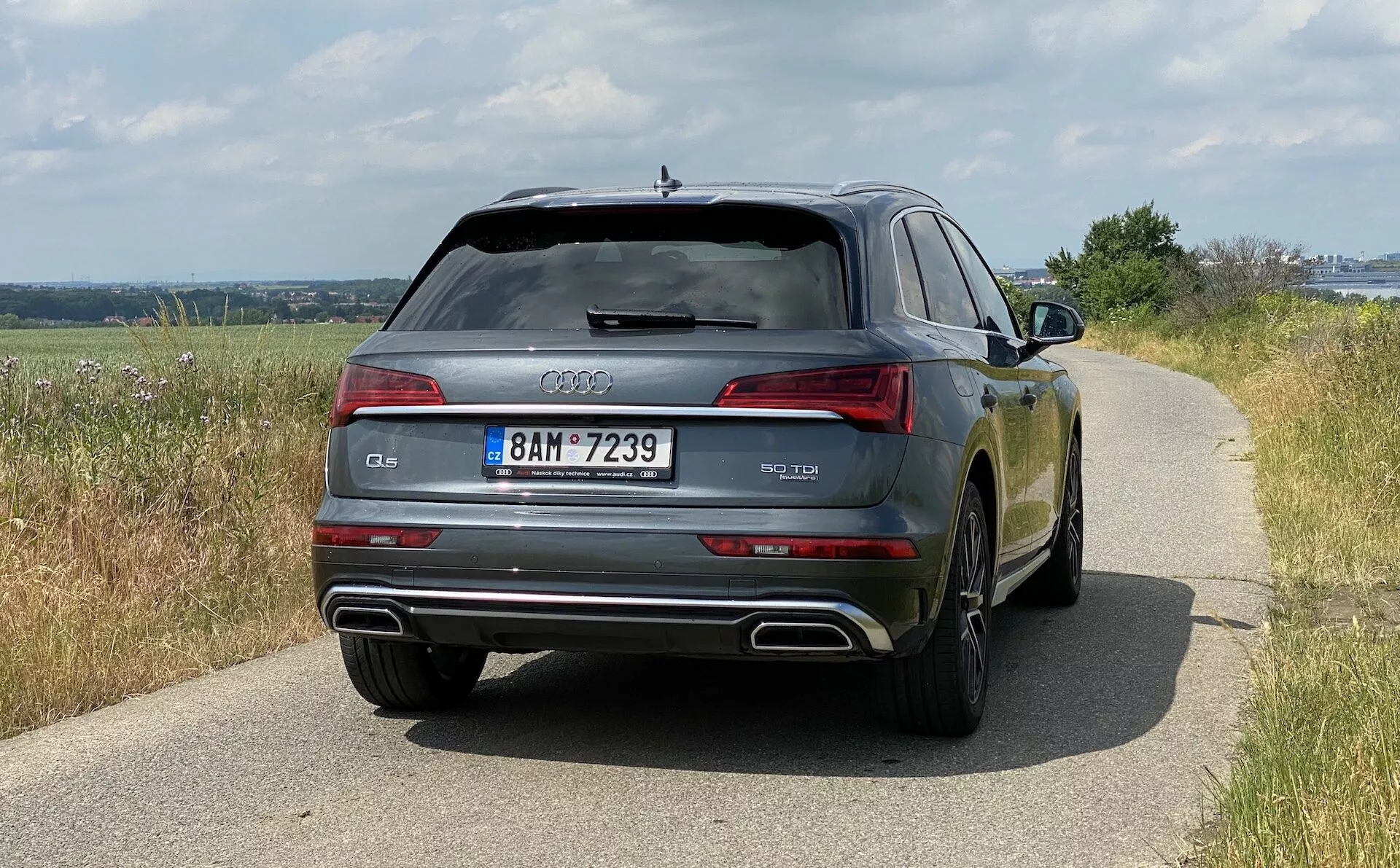 Vue arrière d'une Audi Q5 Sportback de 2020 montrant la largeur du véhicule, y compris les feux arrière, la lunette arrière et les doubles tuyaux d'échappement. La voiture, située sur une route en plein jour montrant un environnement rural, présente une finition de peinture brillante reflétant l'environnement. Les détails de design comme les anneaux Audi, la désignation du modèle et la plaque d'immatriculation sont clairement visibles. Les caractéristiques technologiques telles que les feux à LED, les capteurs de stationnement et la lunette arrière dégivrante sont également évidentes.