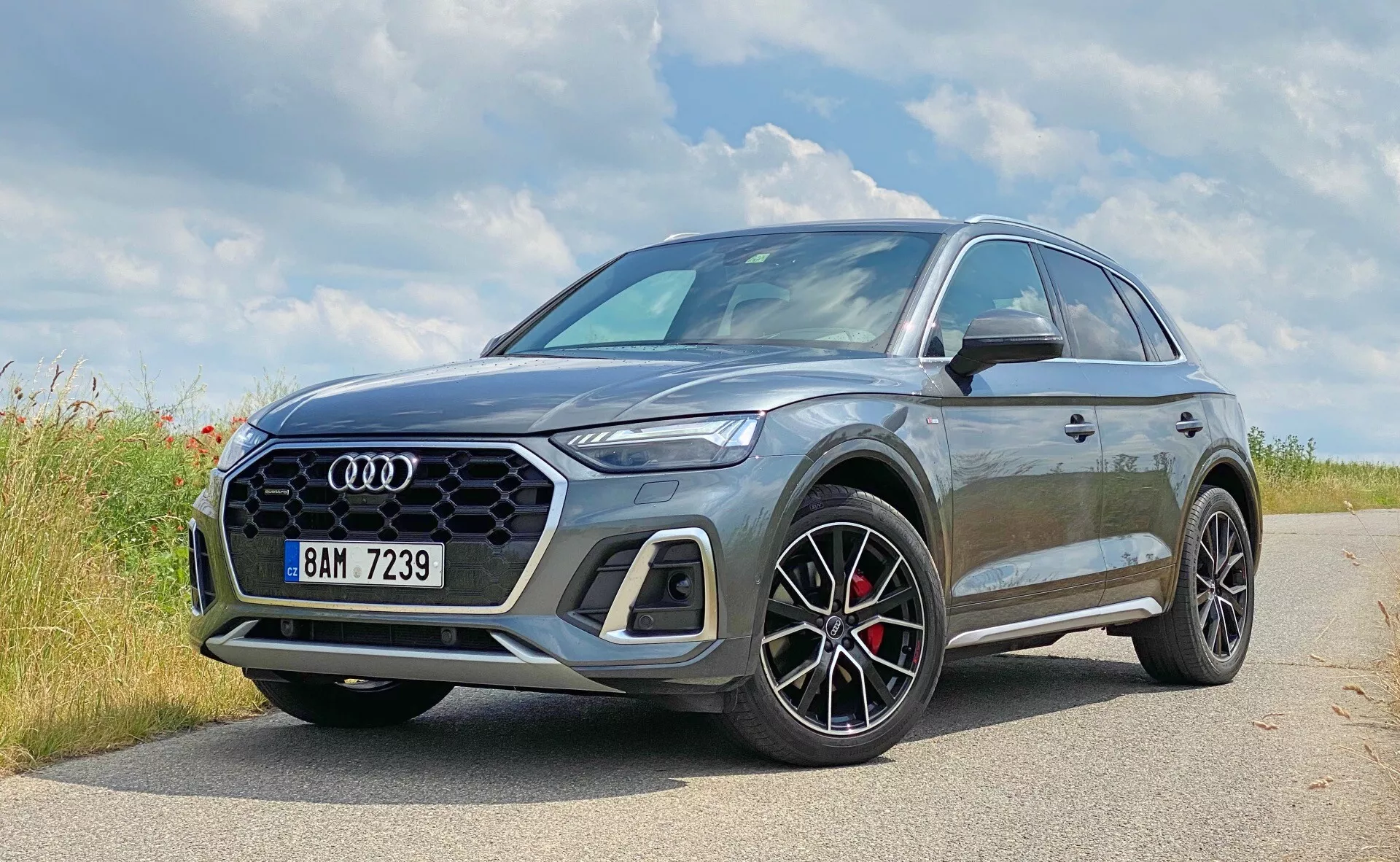 L'image montre un SUV moderne Audi Q5 Sportback 2020 stationné sur une route pavée dans un environnement naturel, possiblement rural. En premier plan, le logo Audi à quatre anneaux est bien en évidence. Cette vue rapprochée, en trois-quarts avant, met en valeur le côté avant gauche et le profil partiel du véhicule, soulignant des éléments de design tels que la grille hexagonale à motif en maille, les phares LED angulaires et les grandes roues à rayons multiples avec des détails noirs et des étriers de frein rouges. La peinture grise métallisée reflète la lumière pour améliorer les contours du véhicule. La garde au sol légèrement augmentée du SUV suggère une polyvalence et des capacités légères tout-terrain, et les rétroviseurs présentent une couleur contrastante indiquant une finition sportive. L'image capture le design automobile de luxe et sportif avec des technologies avancées potentielles non visibles.